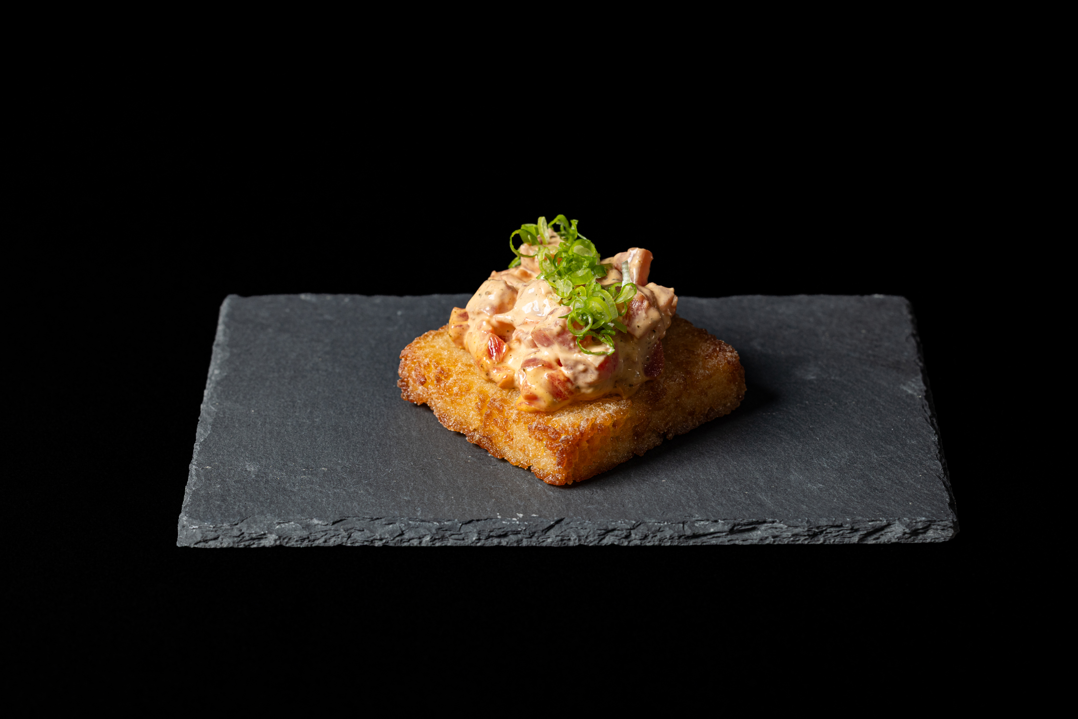 A small piece of fried food topped with a creamy mixture and green garnish on a black slate plate.