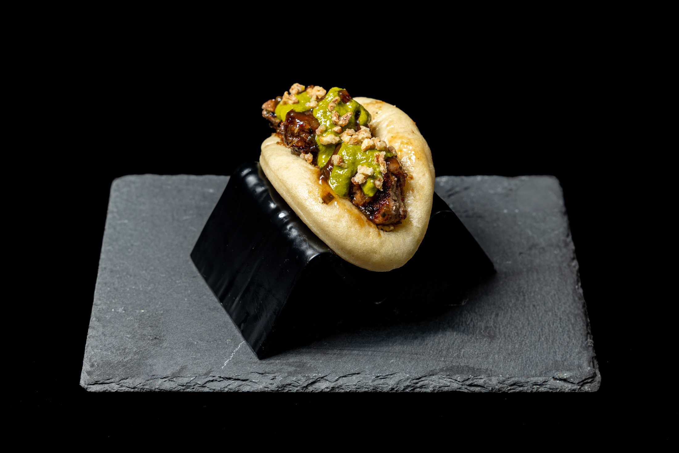 Mini burger with chopped green toppings on a black slate serving board, against a black background.
