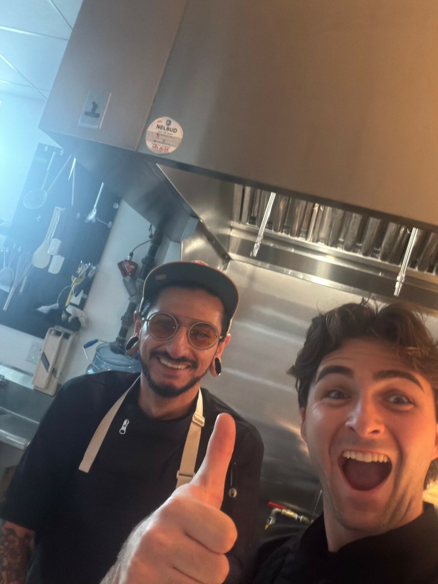 Two young men smiling and taking a selfie in a kitchen, one giving a thumbs-up gesture.
