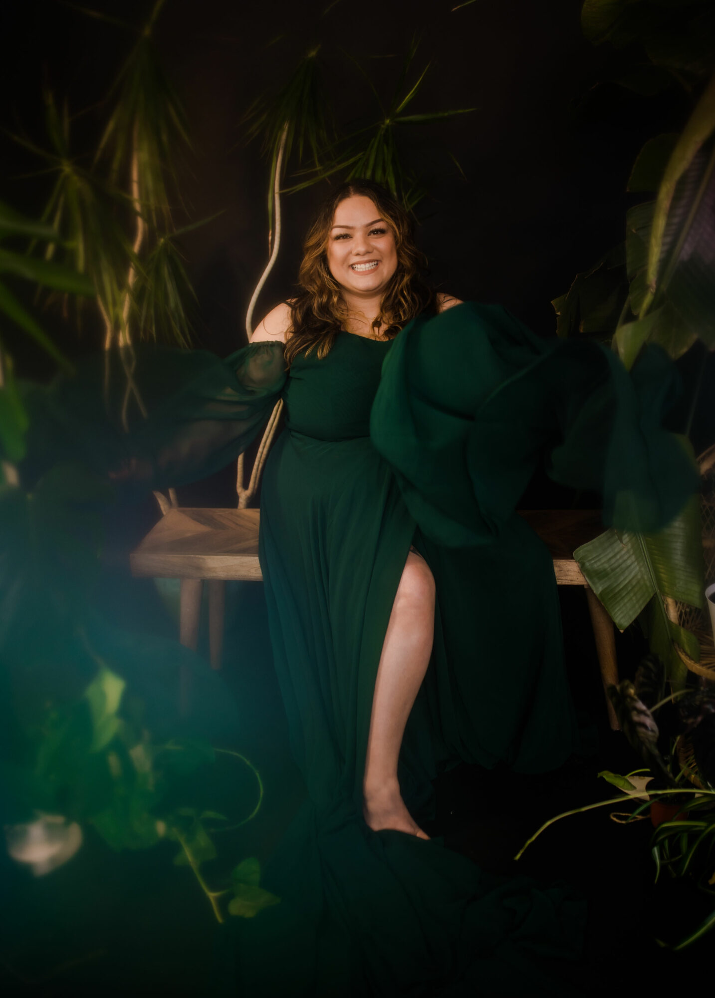 Smiling woman in a dark green dress with a slit, surrounded by plants, sitting on a bench in a garden setting.