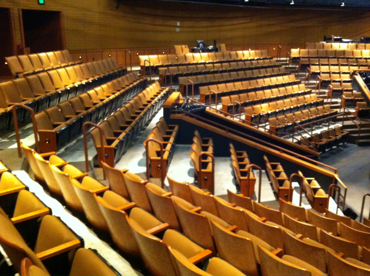 Empty auditorium with rows of yellow seats arranged in a curved pattern, wooden walls, and tiered seating.