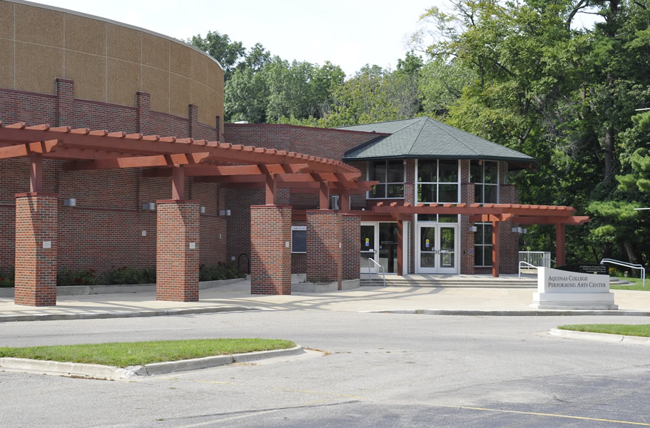 Building with brick walls, large glass entrance, and a wooden pergola structure in front, surrounded by trees and greenery.