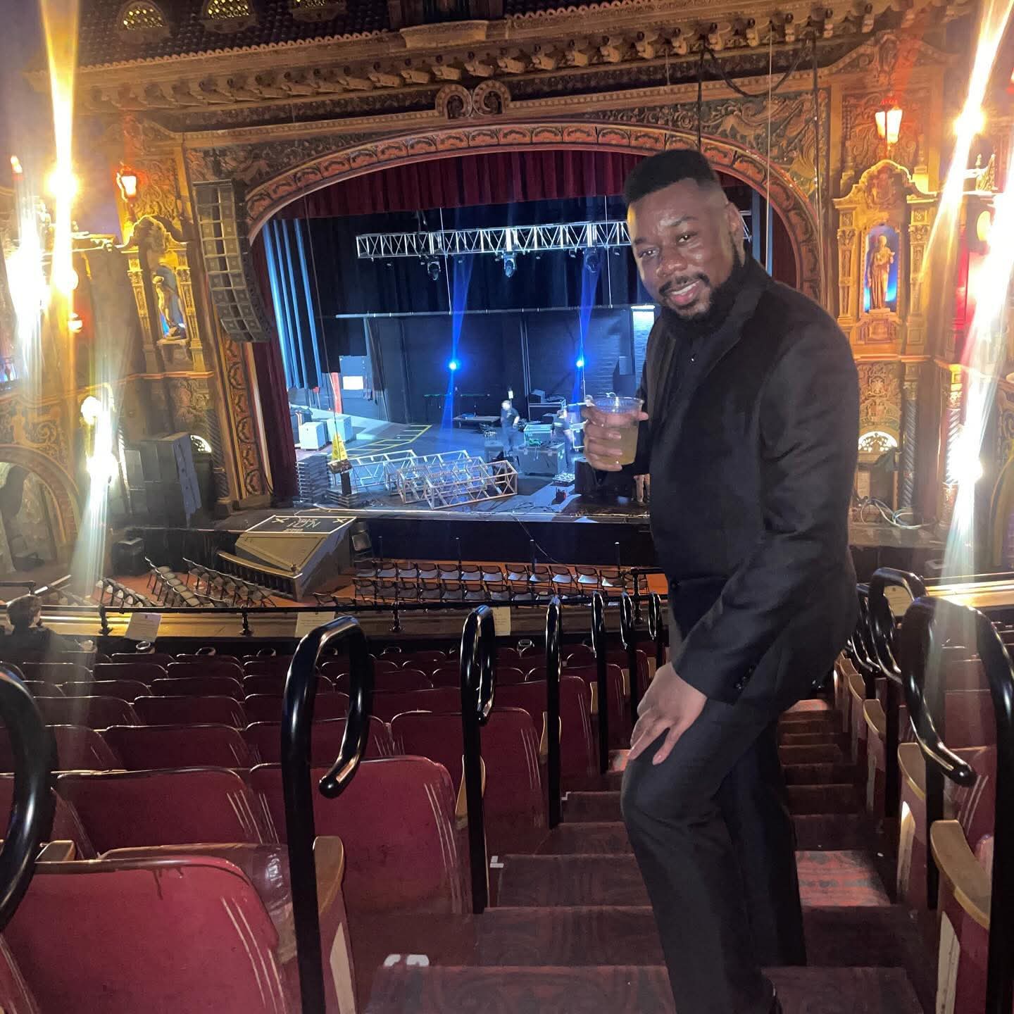 Man in black suit holding a drink, standing in theater balcony with stage in background.