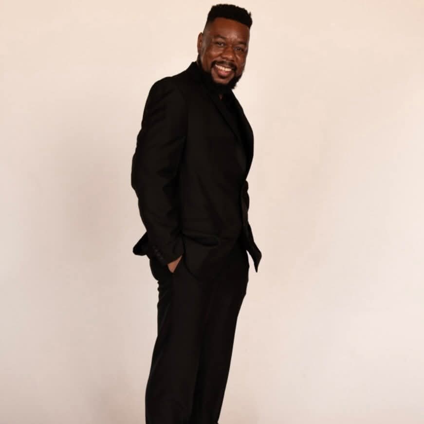 Man in black suit smiling with hands in pockets, standing against a plain background.