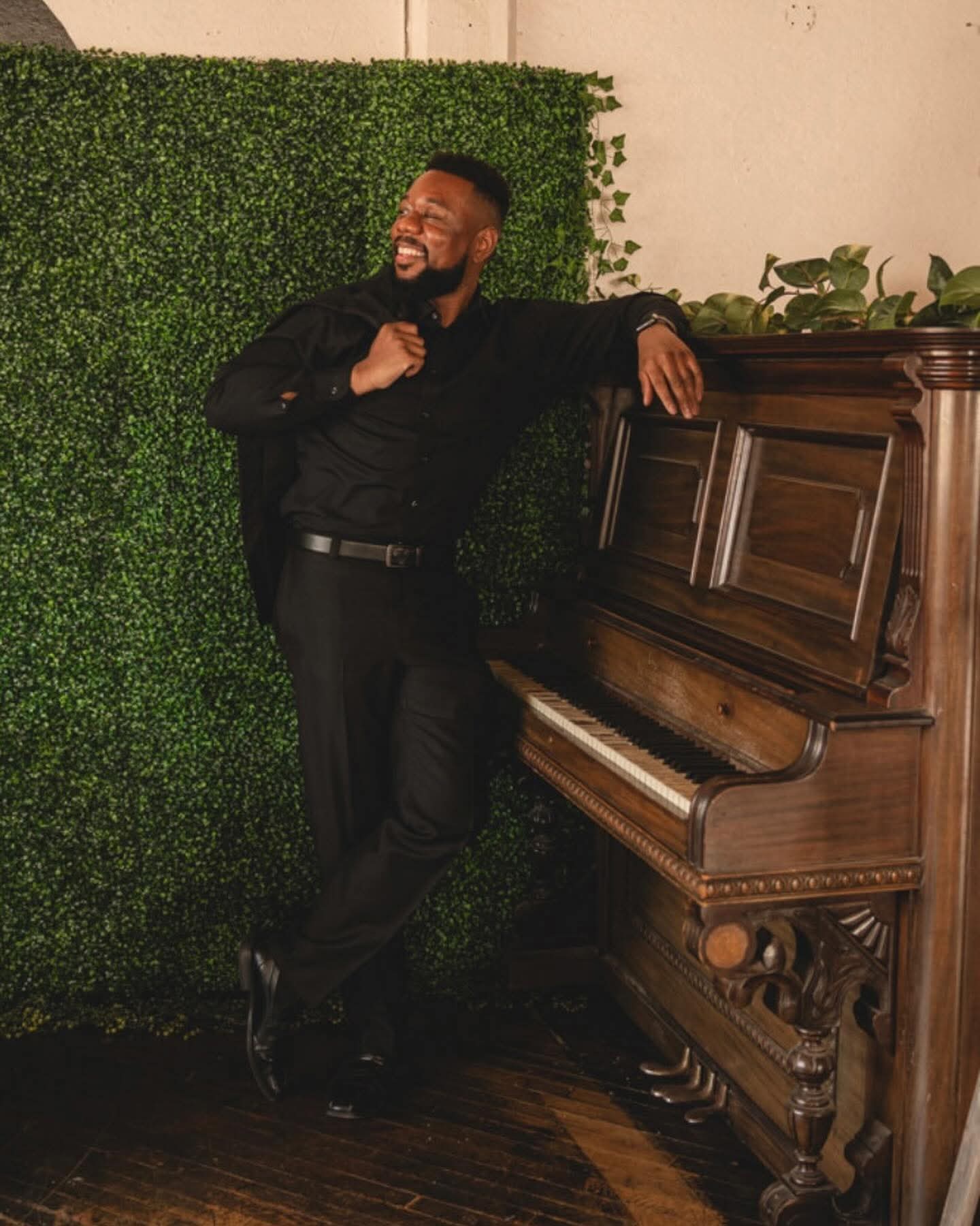 Man in black suit laughing and leaning on a wooden piano, standing in front of a green hedge wall.