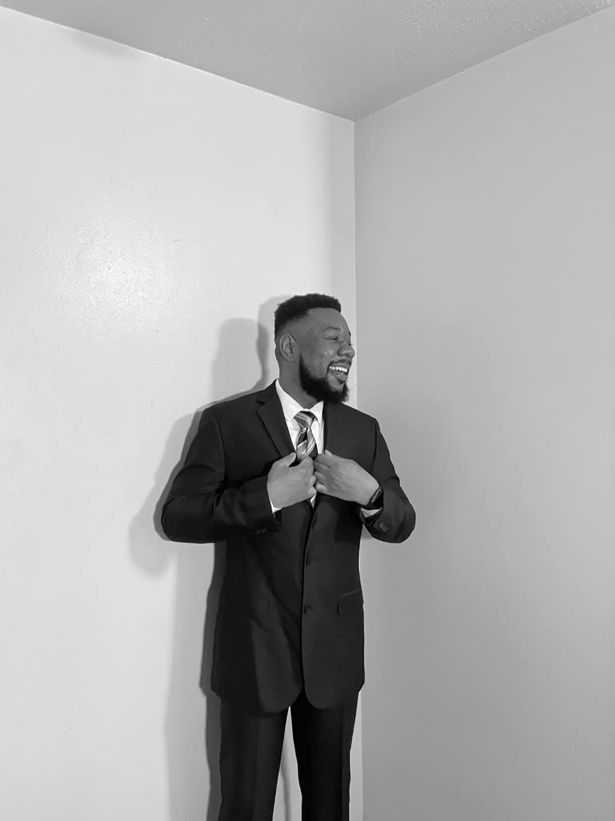 Man in a suit adjusting his tie, standing against a plain wall, smiling, with a shadow on the wall.