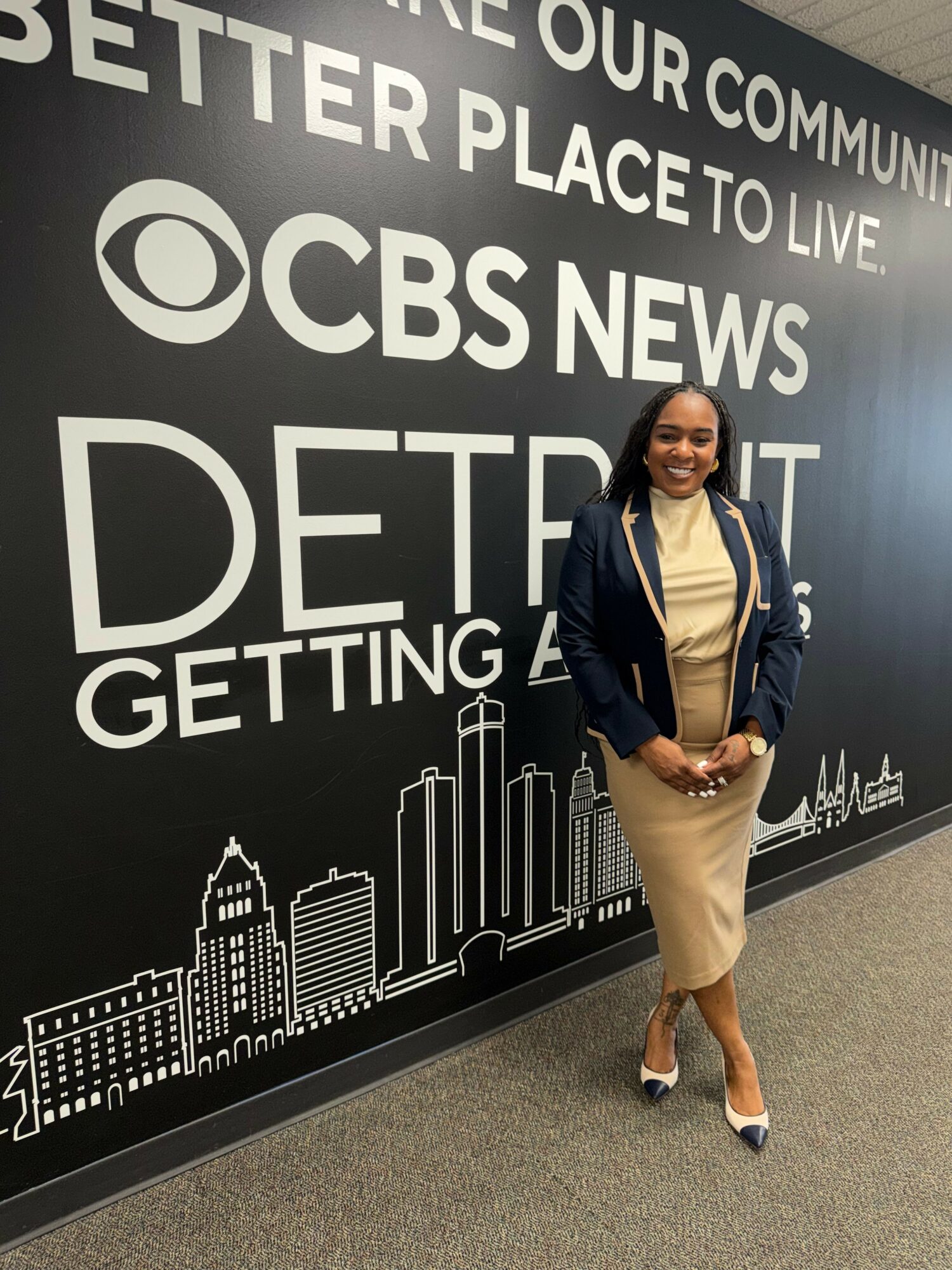 Person standing in front of CBS News display, wearing a dark blazer, beige pants, and white shoes, holding a phone.