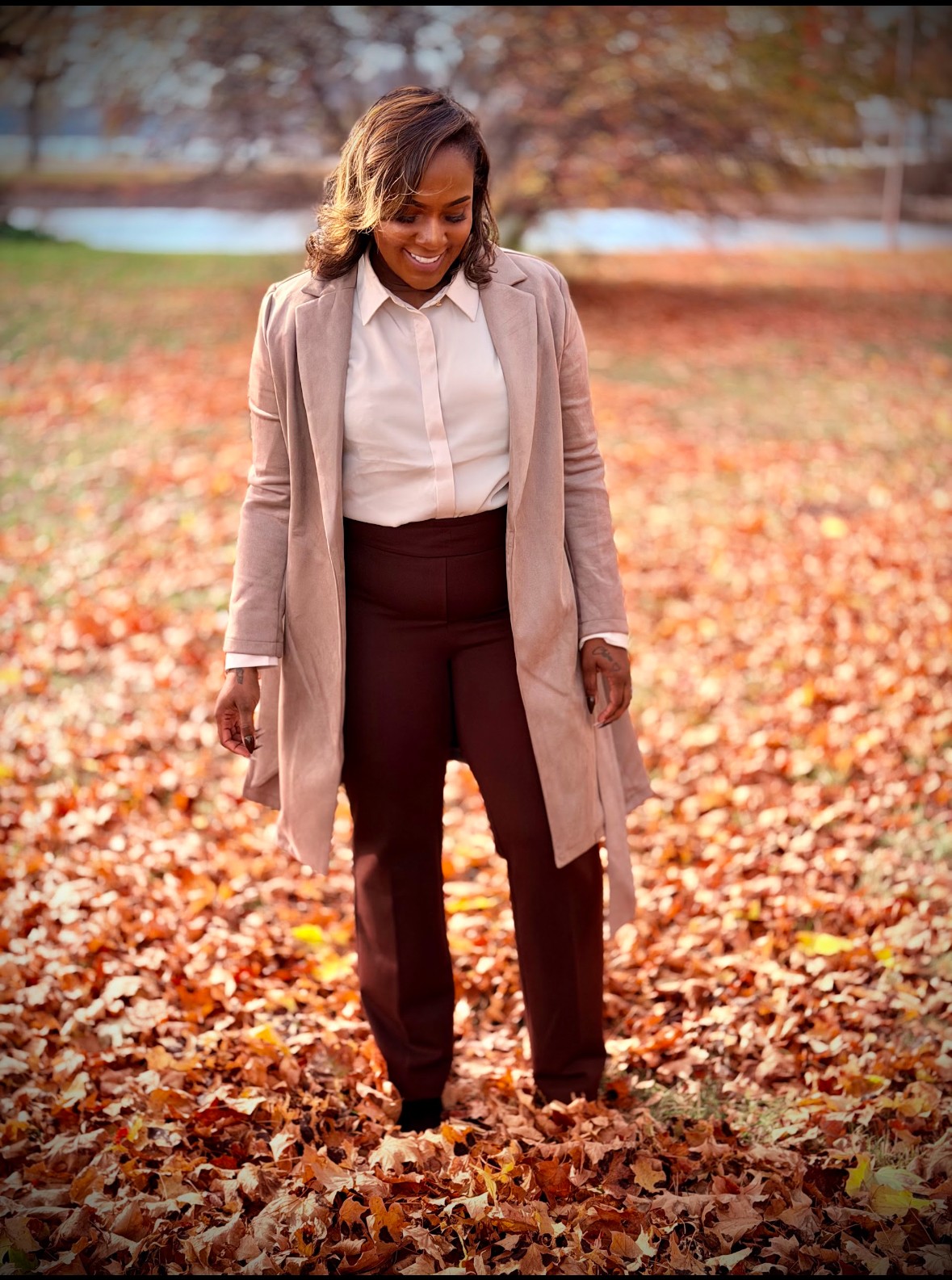 Woman walking through a park covered in fallen autumn leaves, smiling, with trees and a body of water in the background.