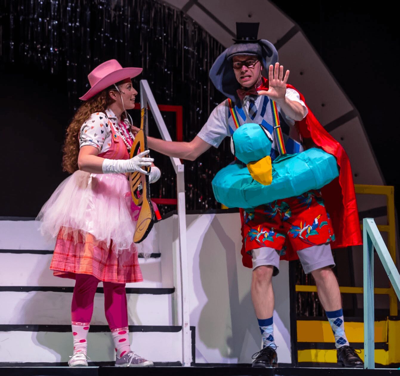 Two performers in colorful costumes on stage, one with a pink hat and the other with a large hat, interacting.