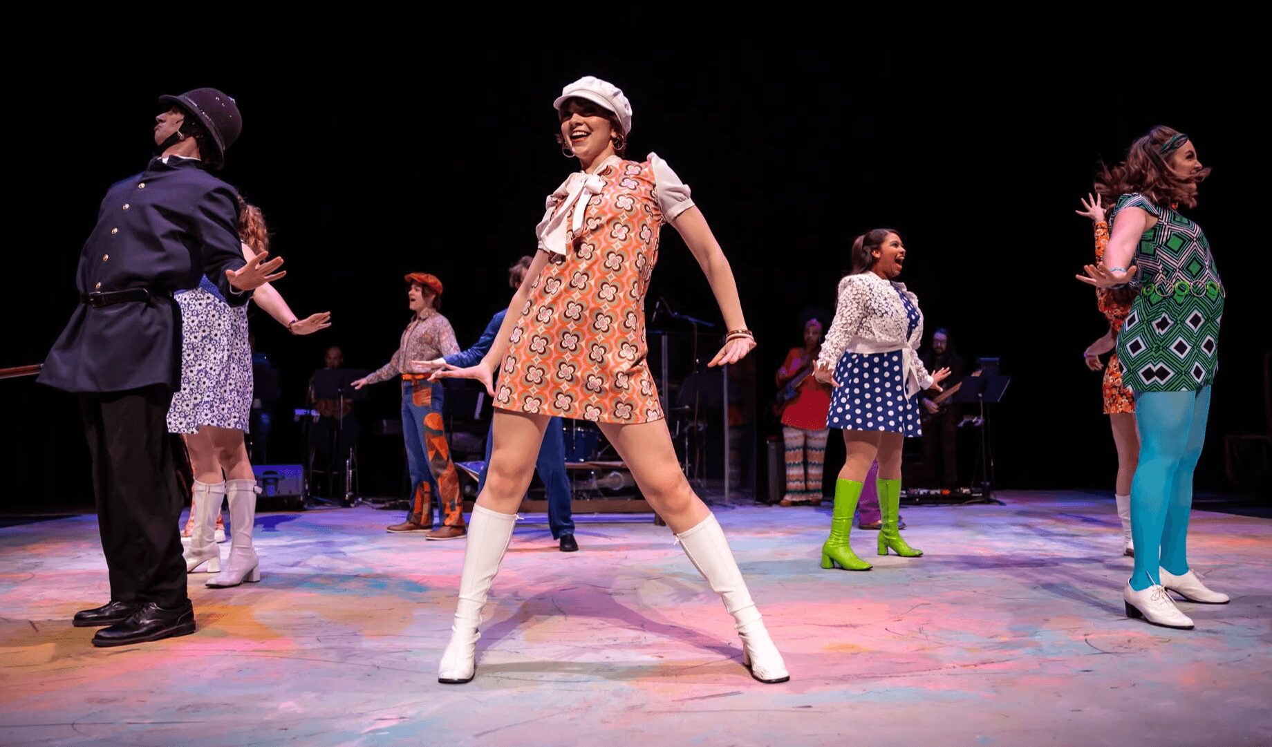 Group of people dancing on stage with colorful lighting, some wearing vintage-style clothing, in a theatrical performance.