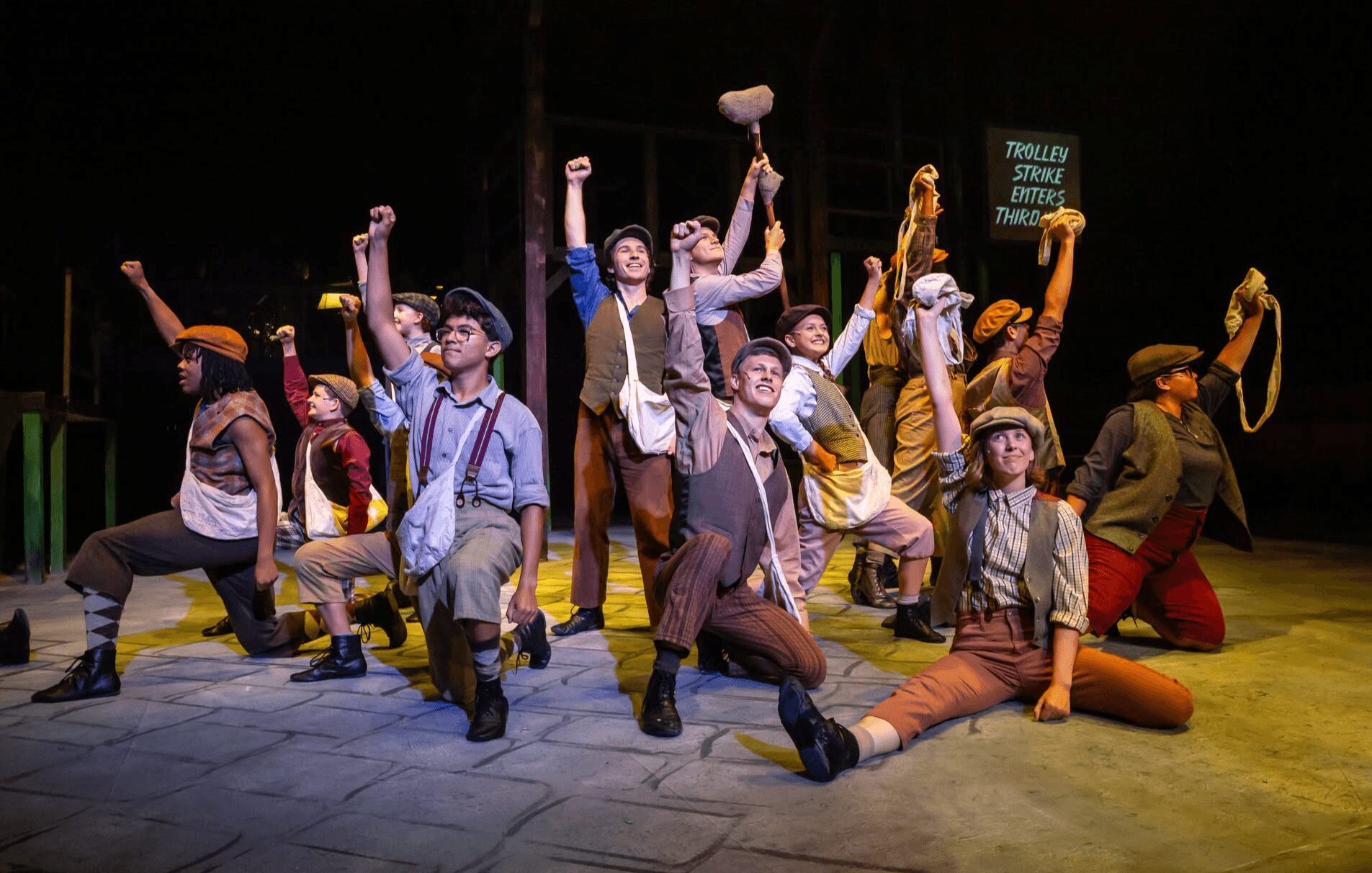 Group of performers in vintage costumes posing on stage with raised fists and hats, some kneeling or sitting.