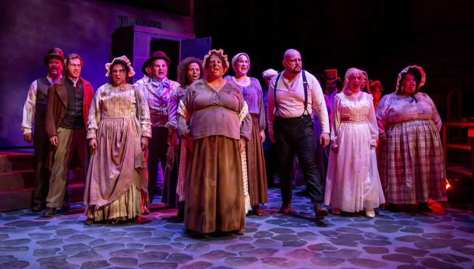 Group of people on stage in period costumes, standing in a line, with a dark background and cobblestone floor.