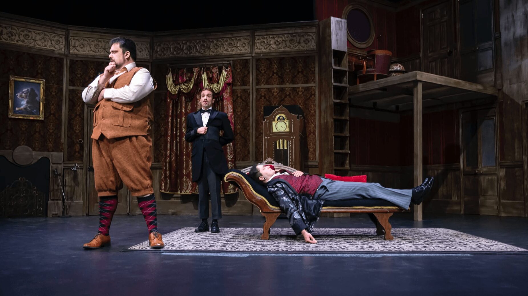 Three actors on stage in a theatrical scene, one lying on a bench, two standing, set in a vintage room.