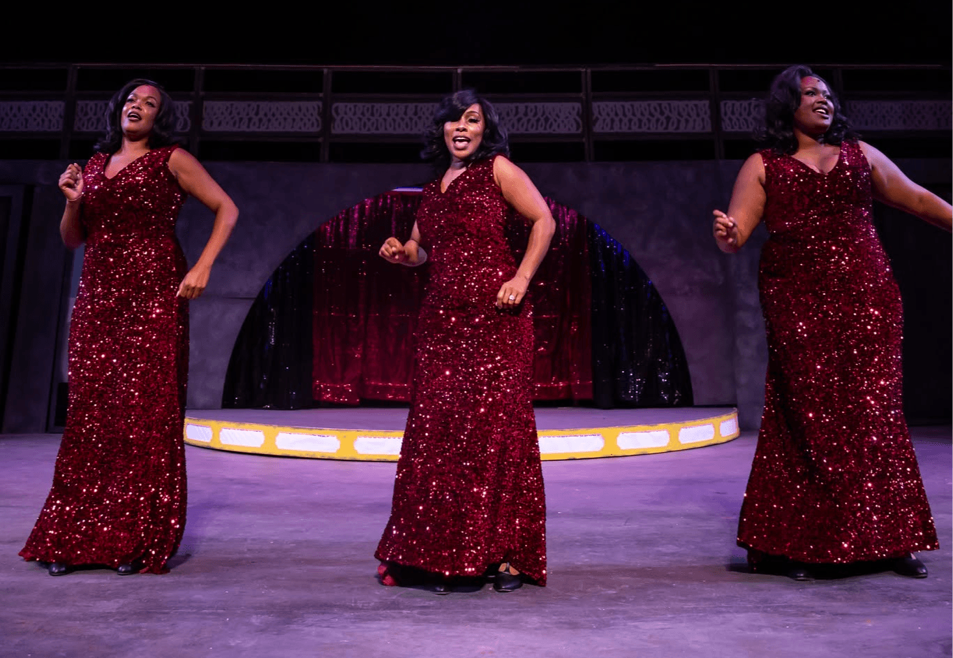 Three women in matching red, sparkly dresses perform on stage with a dark background and curtain.