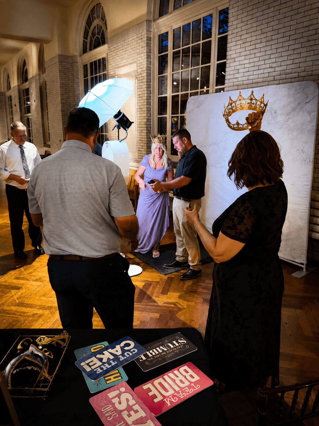 People gather around a woman in a purple dress and a man, with a backdrop featuring a crown logo, in a well-lit room.