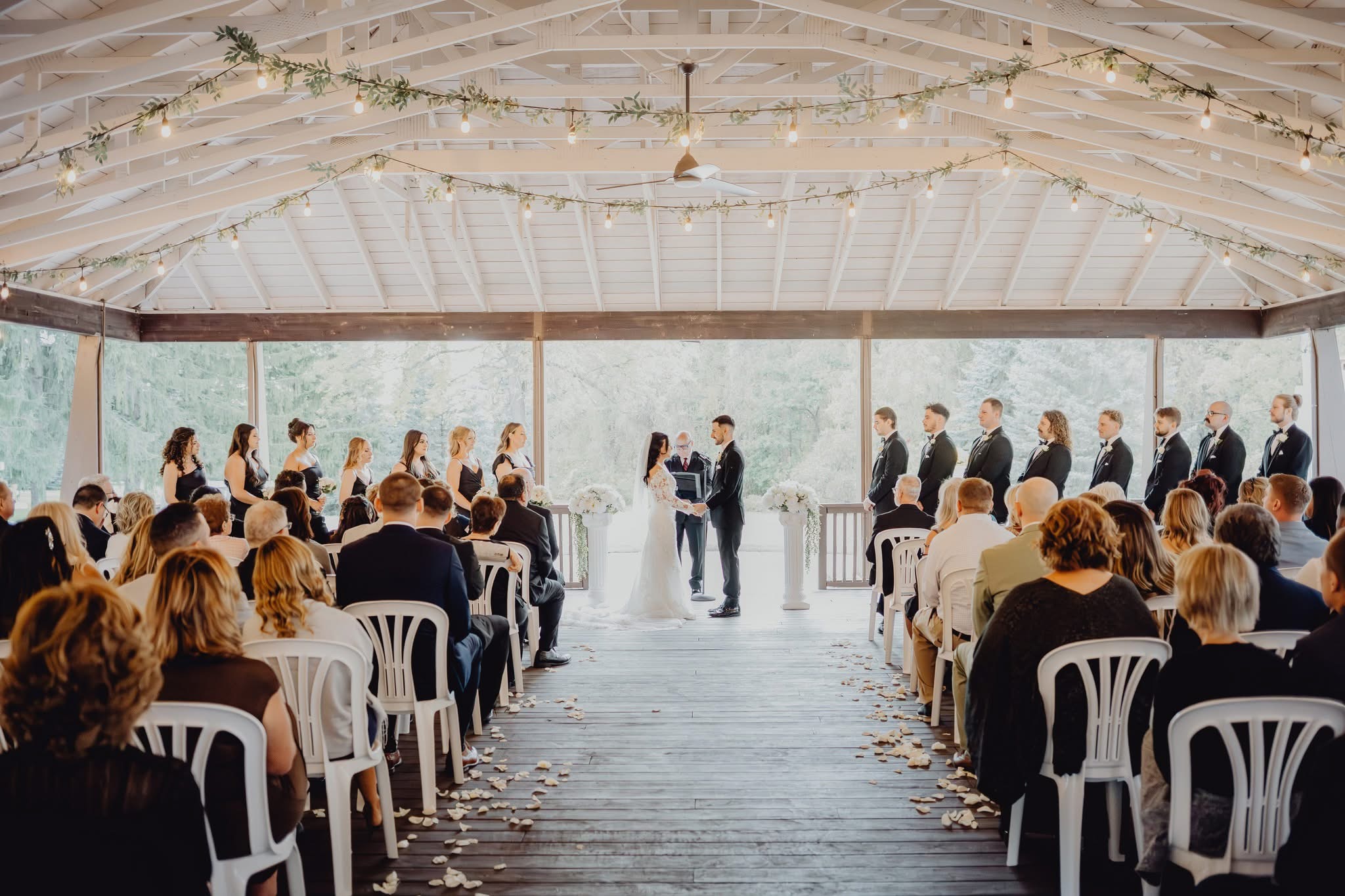 Wedding ceremony with bride and groom exchanging vows under a decorated canopy, guests seated on both sides, bright natural light.
