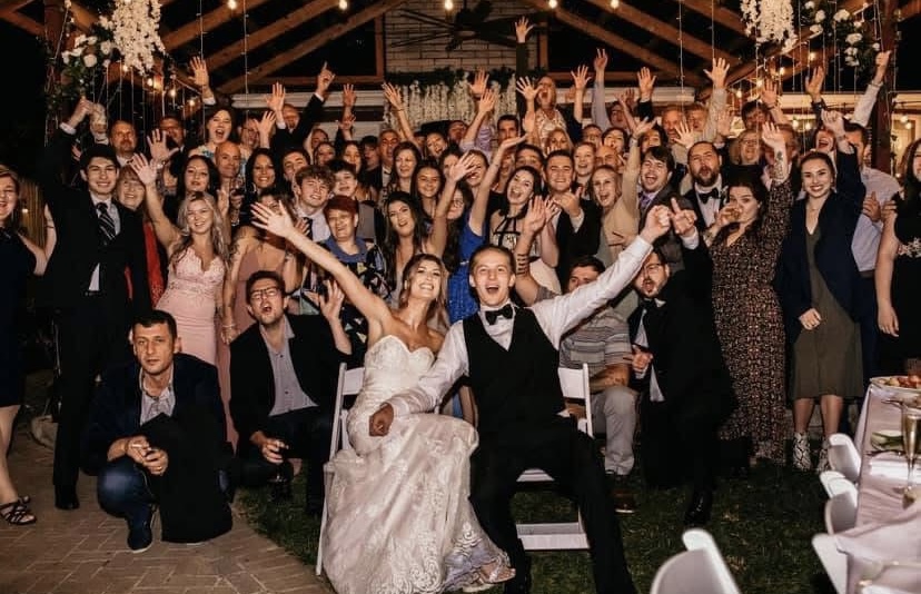 Large group of people celebrating at a wedding reception, with a bride and groom in front, smiling and raising hands.