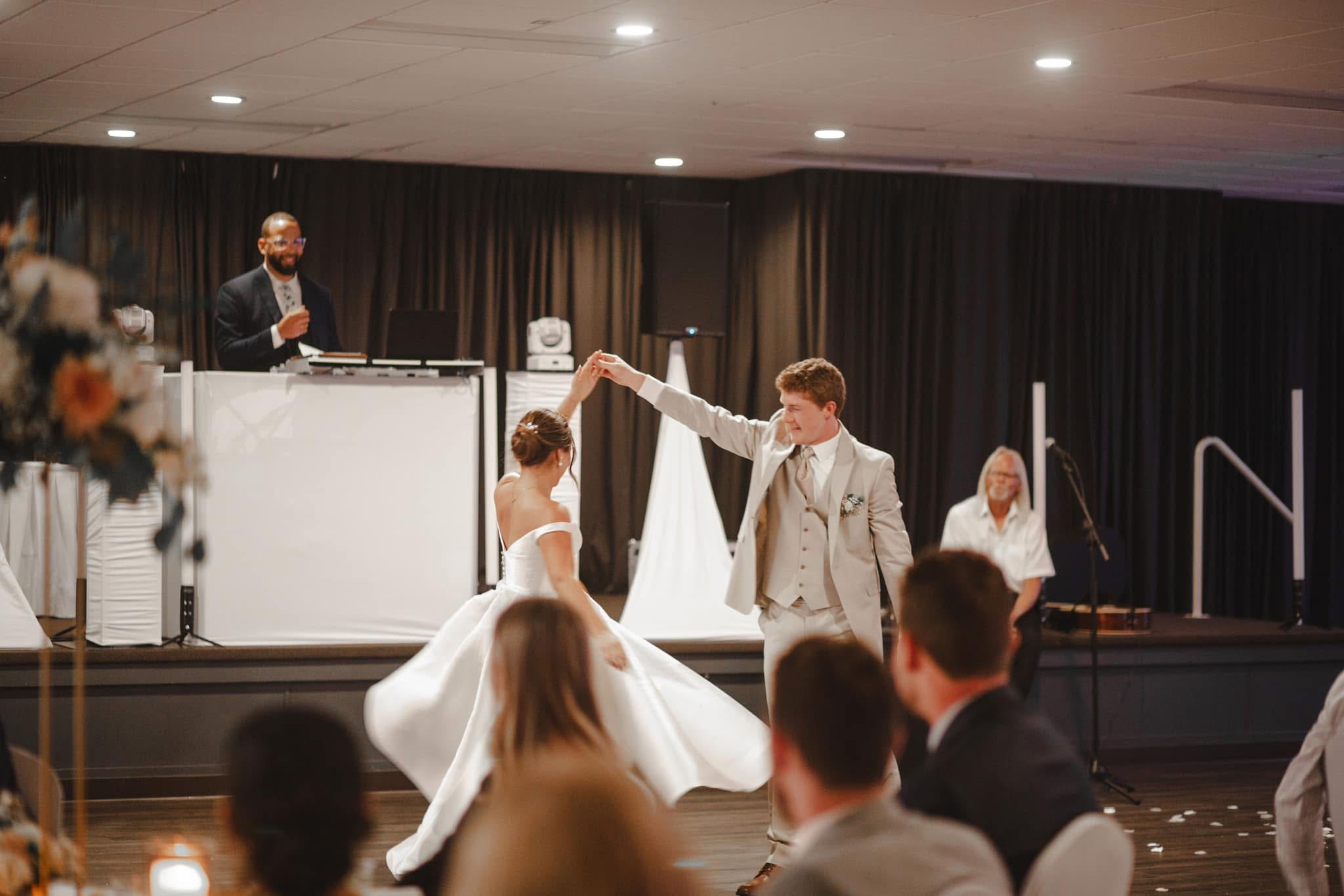 A couple dances at their wedding reception with a DJ and a woman in the background, guests watching, and a stage backdrop.