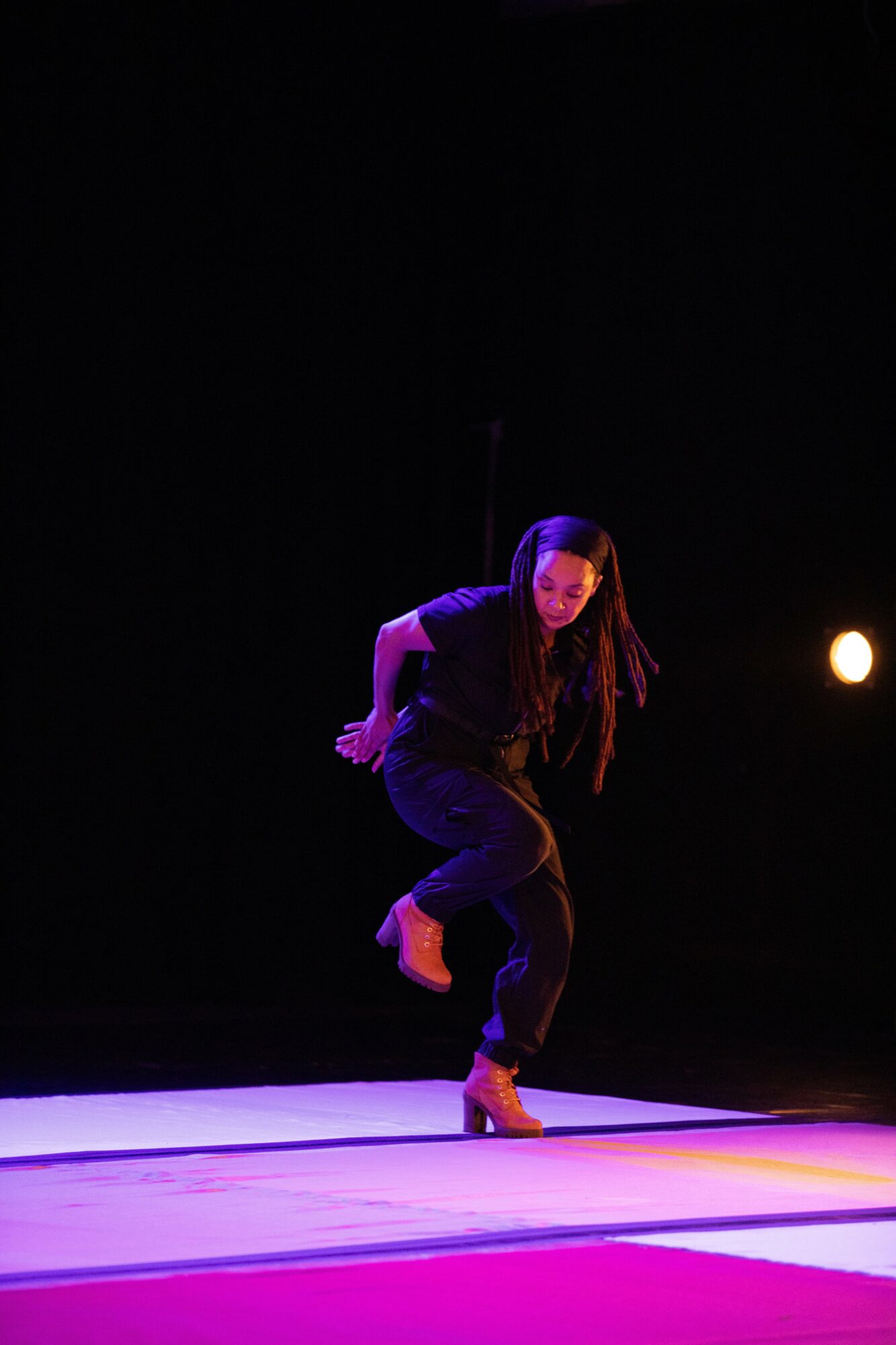 A woman with long hair performs a dance move on stage, illuminated by purple and yellow lighting, with a dark background.