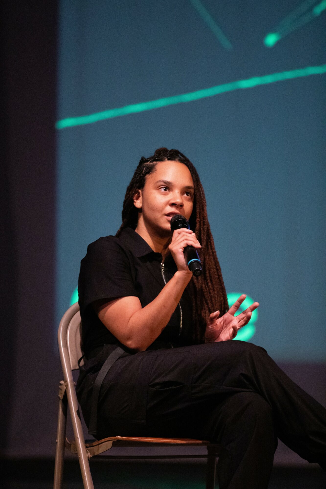 Young woman with long hair speaking into a microphone while sitting on a chair on stage.