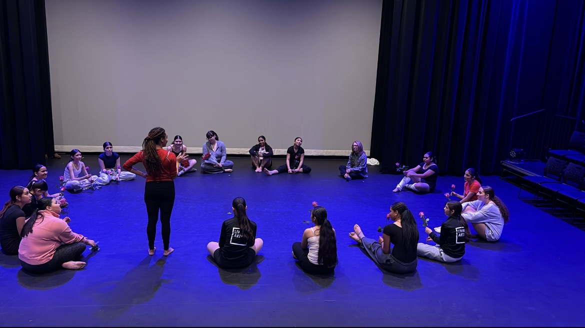 Group of children sitting in a circle on a stage with a woman standing in the center, blue lighting, dark curtains.
