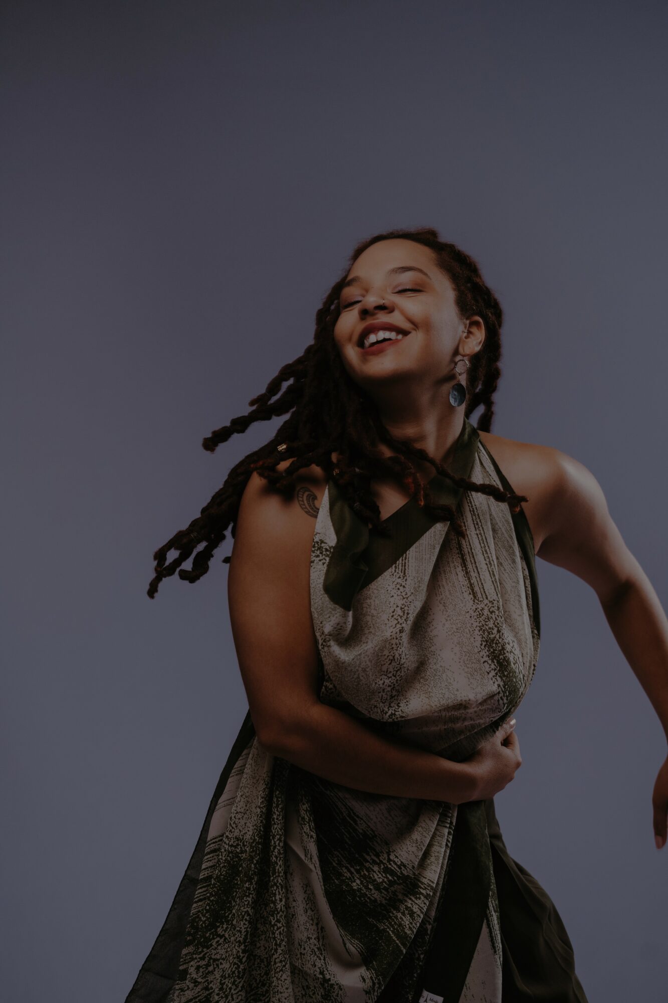Smiling woman with dreadlocks wearing a sleeveless dress, standing against a plain background.