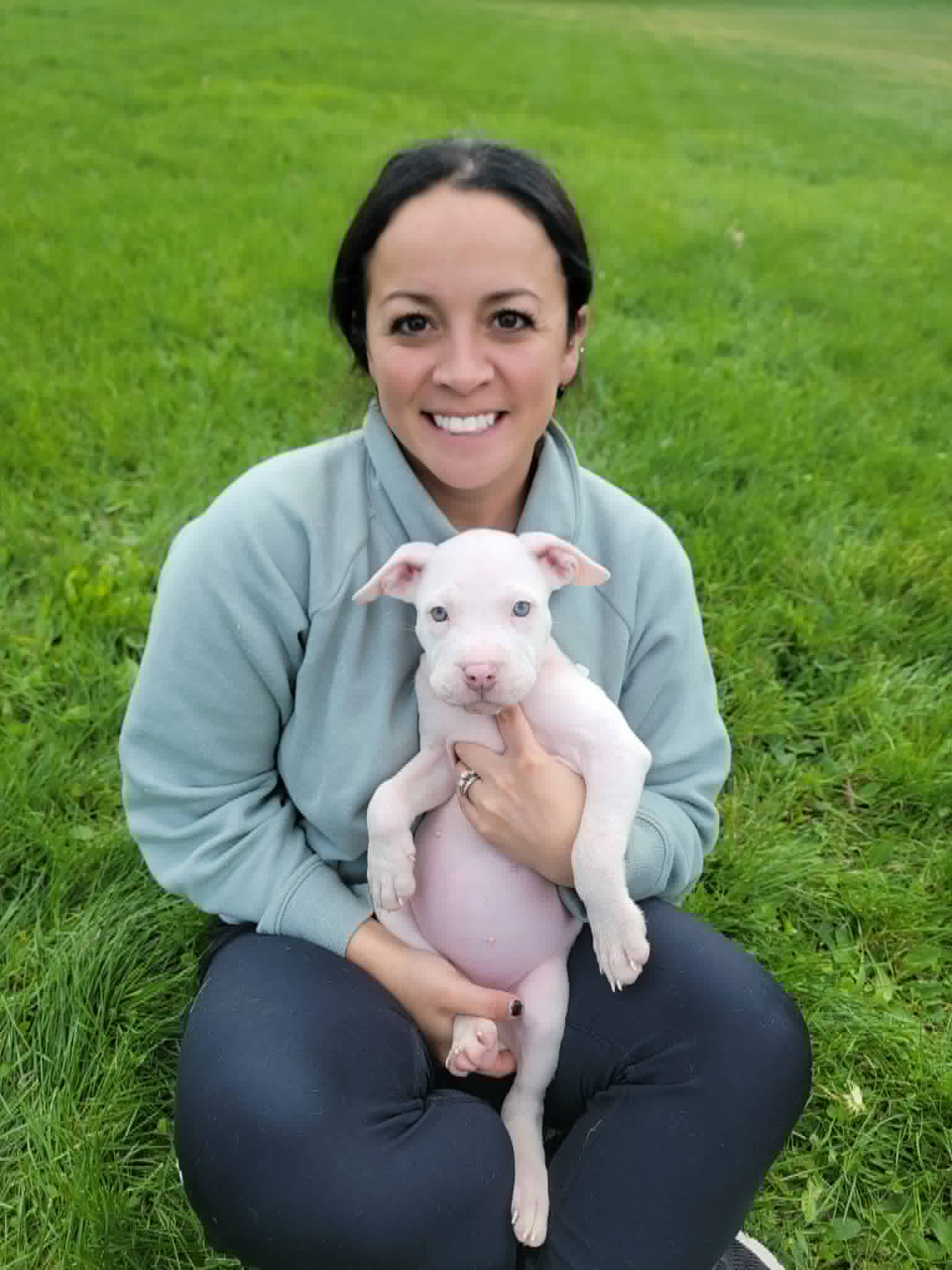 Woman sitting on grass holding a white puppy, smiling, outdoors with green grass background.