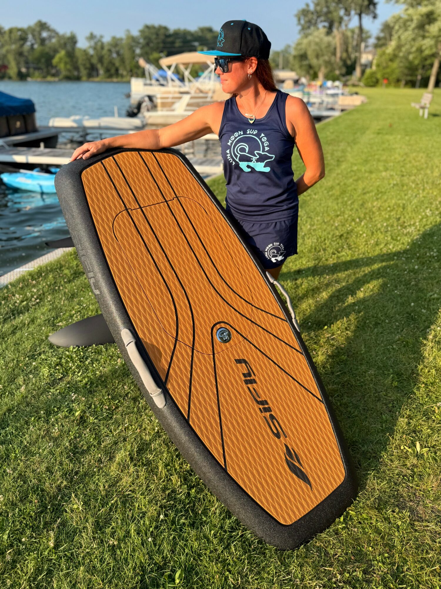 Child in a navy tank top and cap holding a large wooden paddleboard on grass near water with boats in background.