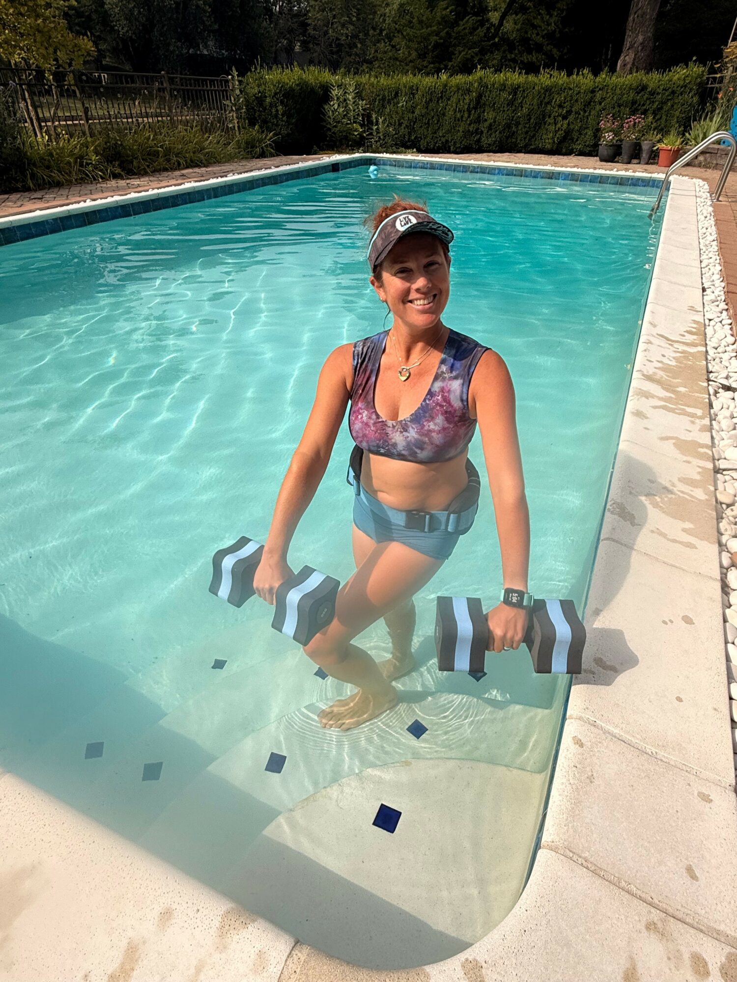 Woman standing in swimming pool holding a striped float, wearing a cap and swimsuit, with trees and a fence in background.
