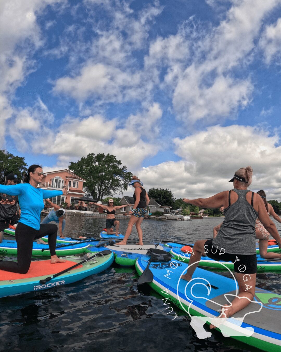 People paddleboarding on water under a partly cloudy sky, with houses and trees in the background.