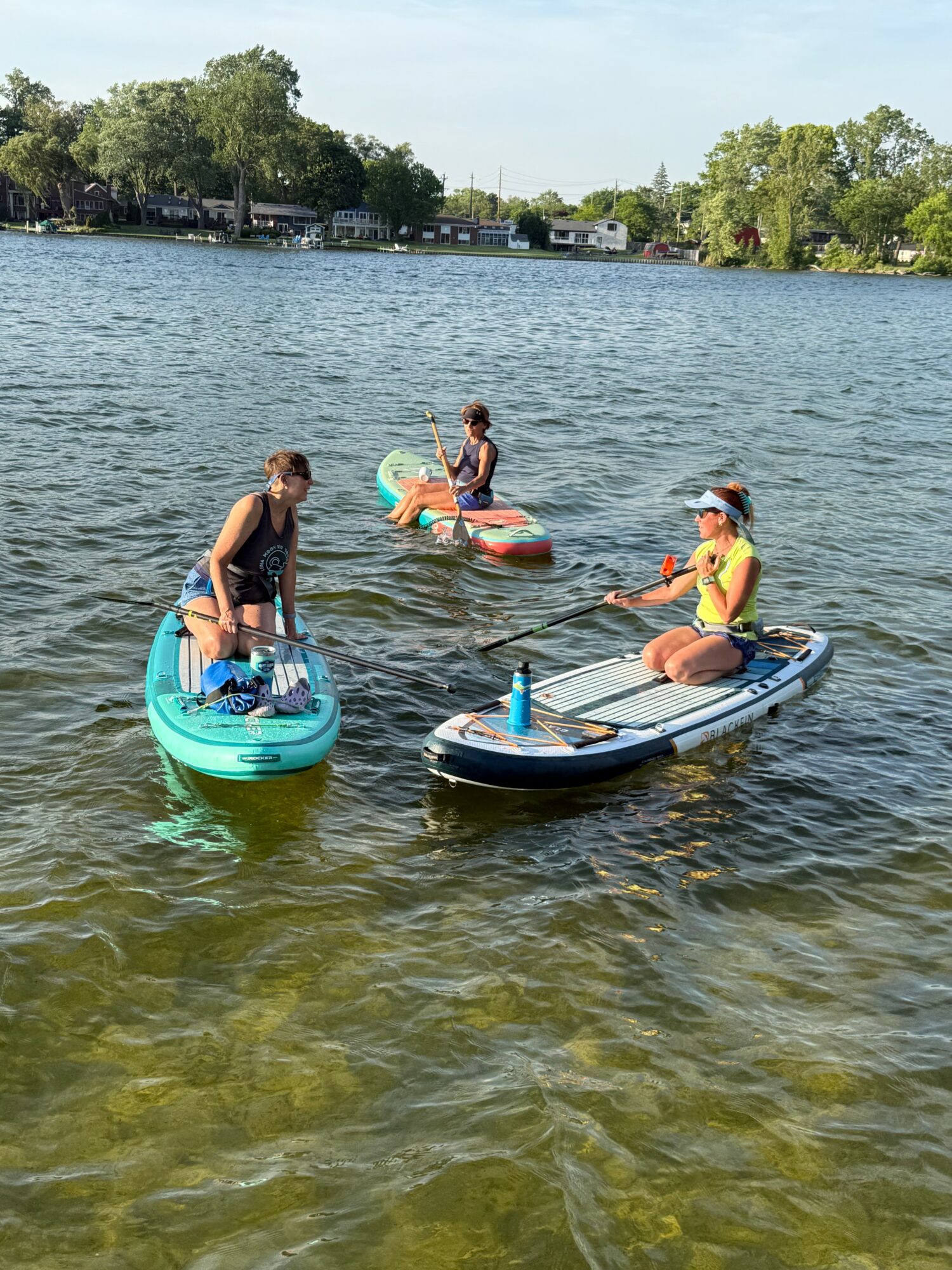 Three people on paddleboards and one person in the water on a lake, with trees and houses in the background.