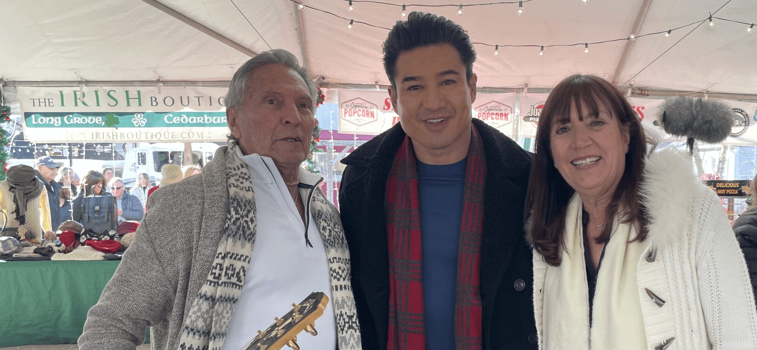 Three people smiling at an outdoor event under a tent, with market stalls in the background.
