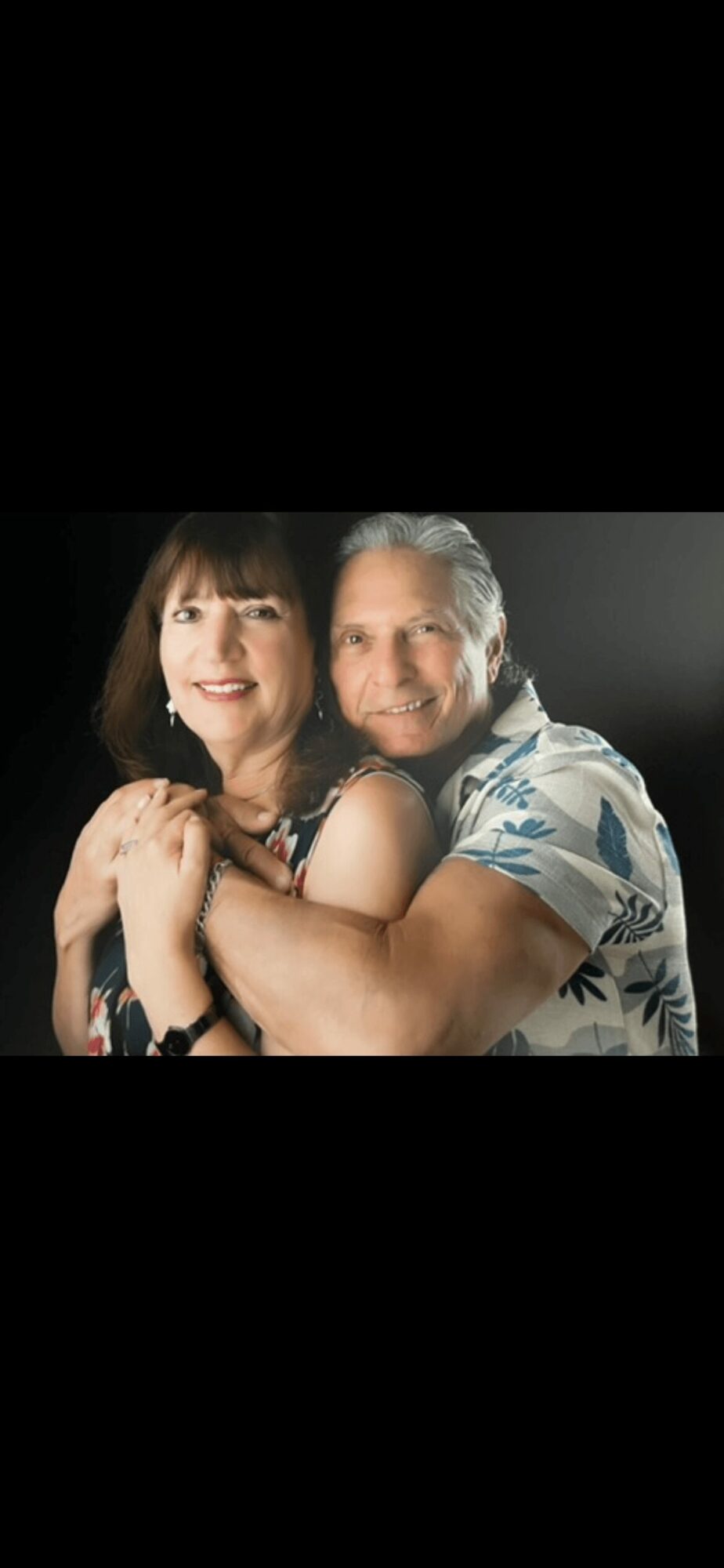 A smiling couple embraces each other, woman with dark hair and man with gray hair, against a dark background.