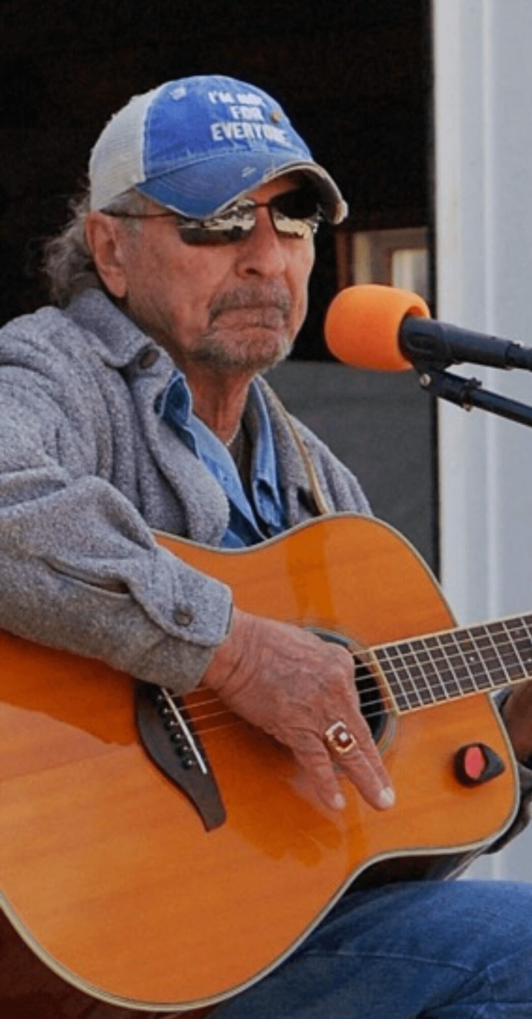 Man wearing sunglasses and a cap playing an acoustic guitar outdoors, with a microphone nearby.