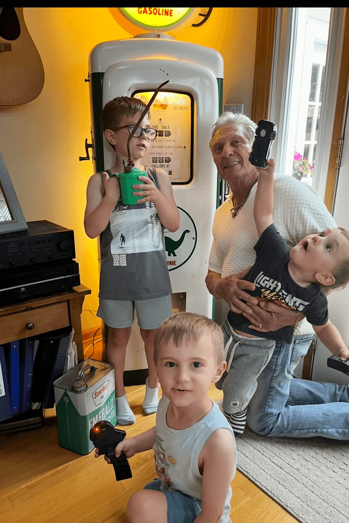 Two children and an elderly woman in a room with a vintage gas pump, one child holding a toy gun, smiling.