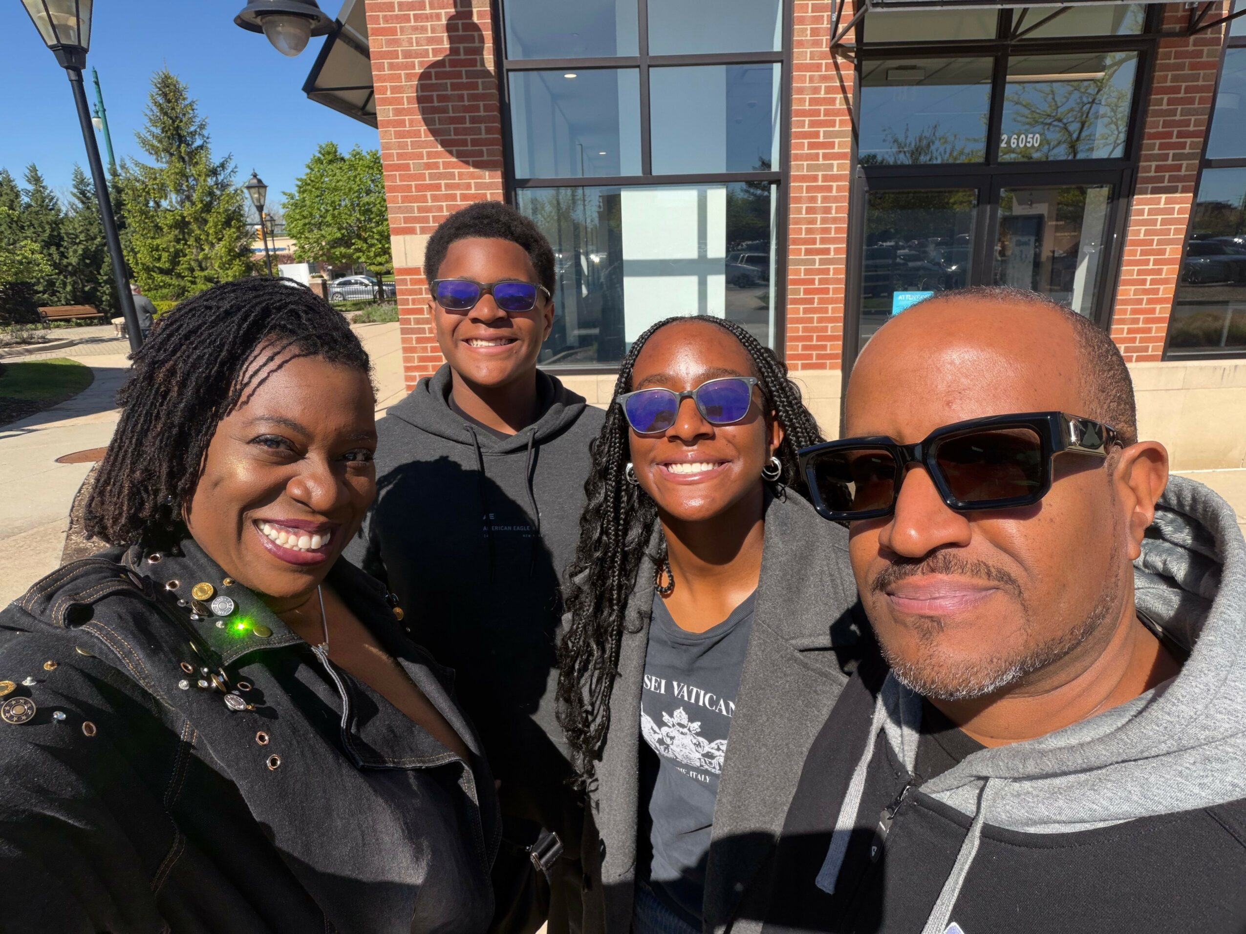 Four people smiling outdoors in front of a building with large windows, sunny day, some wearing sunglasses.