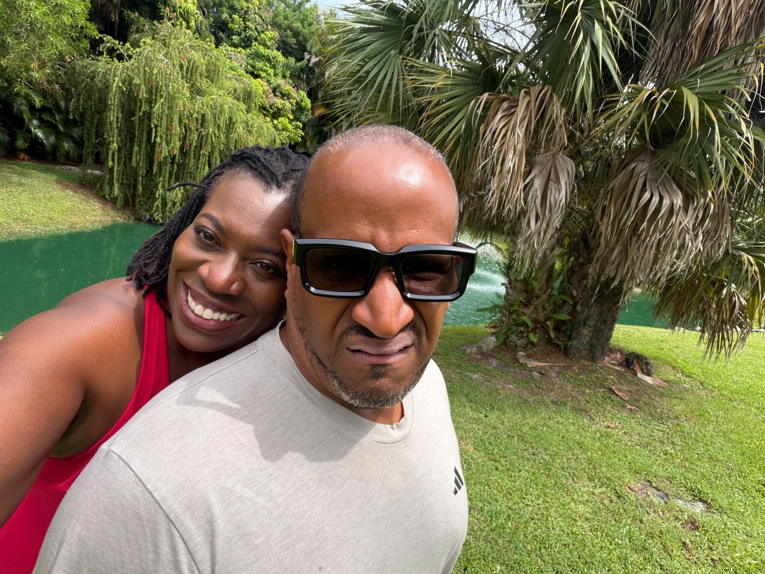 Two people smiling outdoors near a tree and a pond, one wearing sunglasses and a white shirt, the other in a red top.