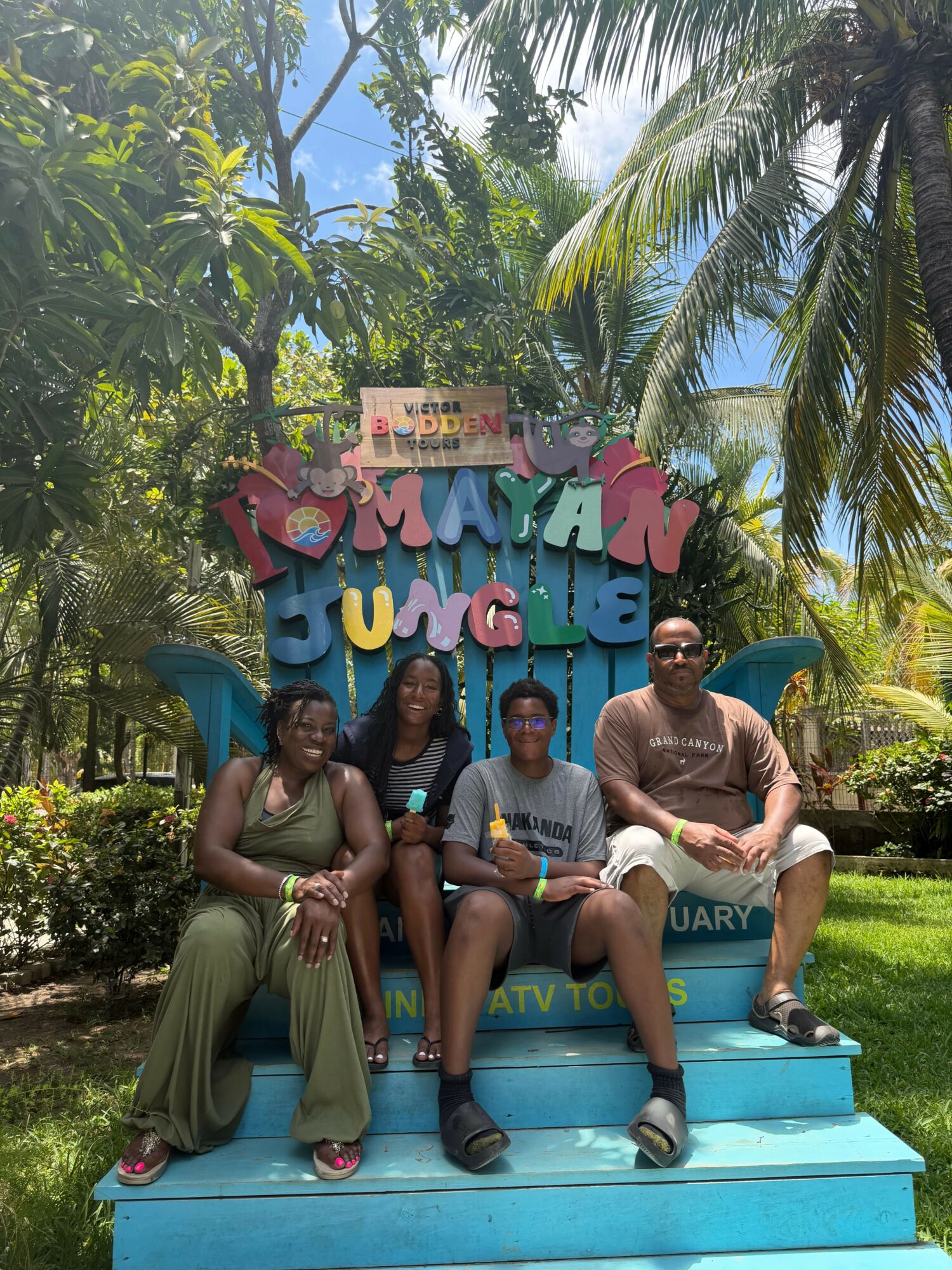 Three people sitting on a blue platform in a tropical garden with palm trees and greenery, smiling and posing for the photo.