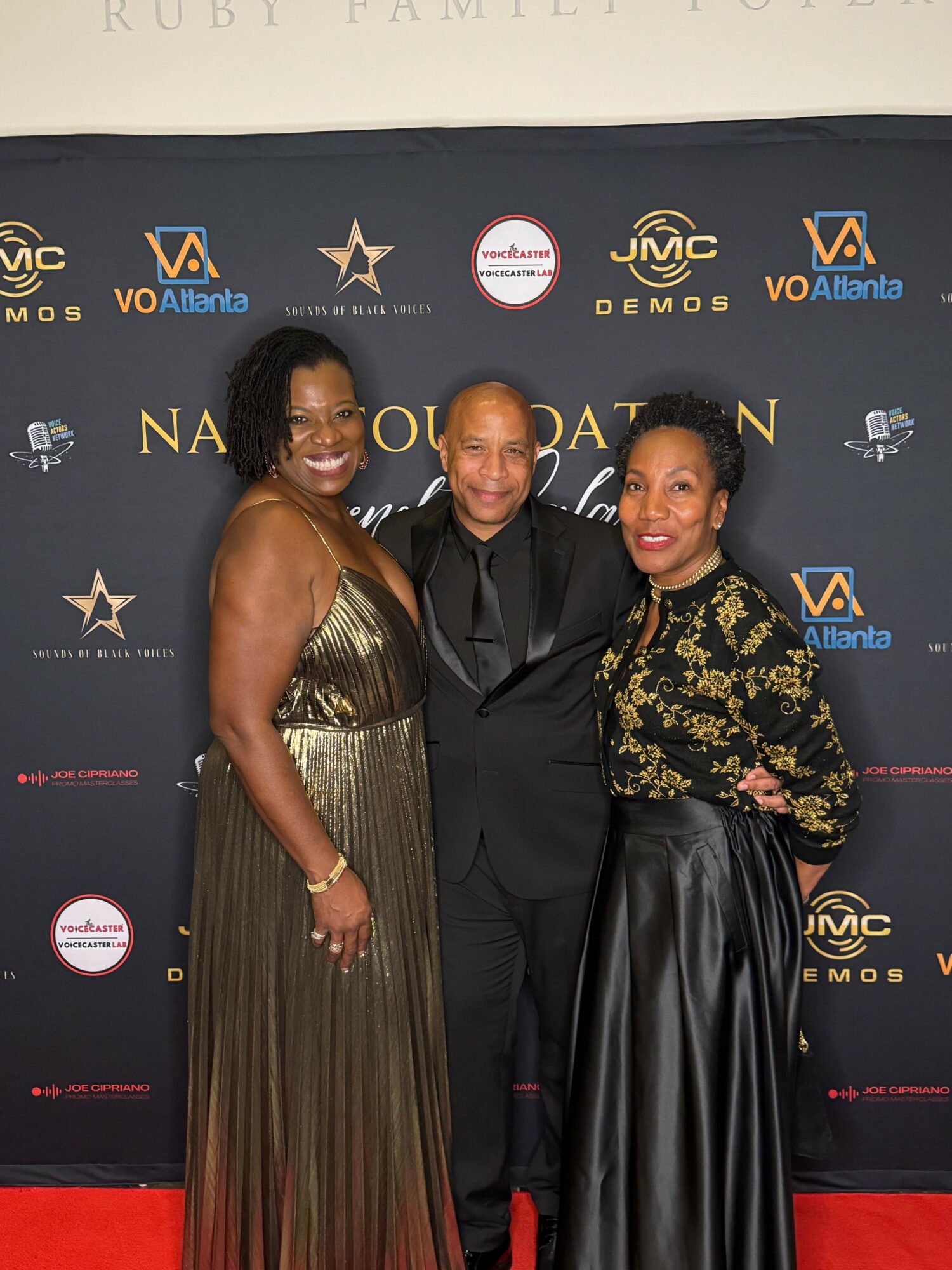 Three people standing together at an event, posing in front of a backdrop with logos and text, smiling at the camera.