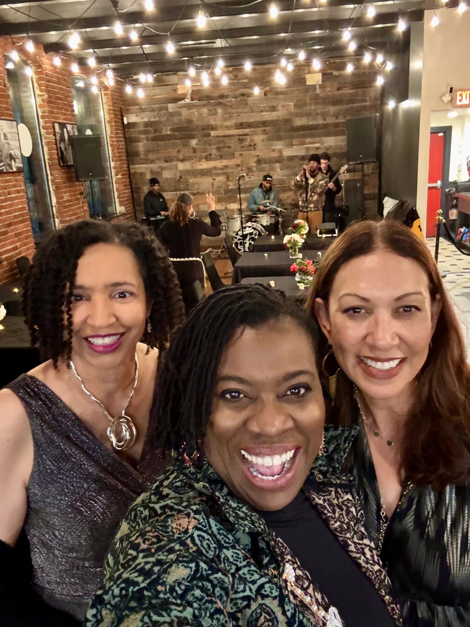 Three women smiling in a decorated indoor venue with string lights and a band performing in the background.