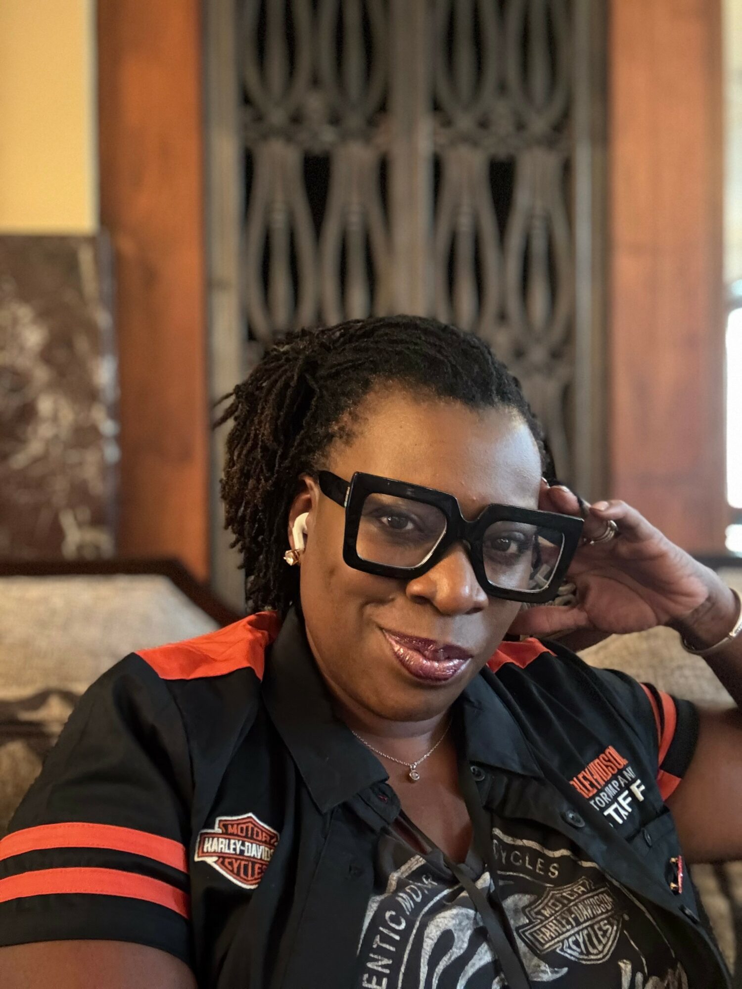 Woman with glasses and earbuds, wearing a black shirt with red accents, sitting indoors with a decorative background.