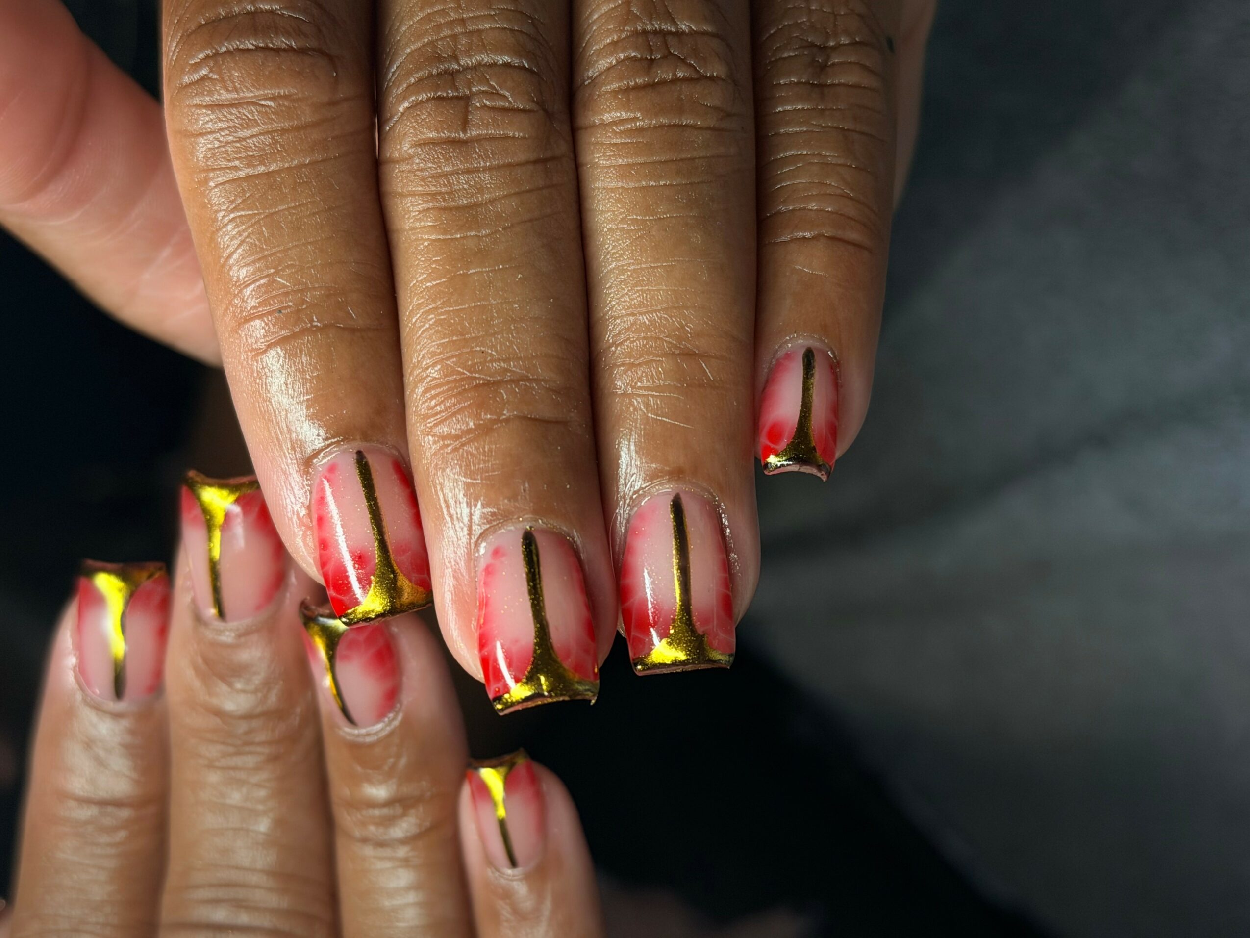 Close-up of hands with decorated nails featuring red, yellow, and black designs, and gold accents, against a dark background.
