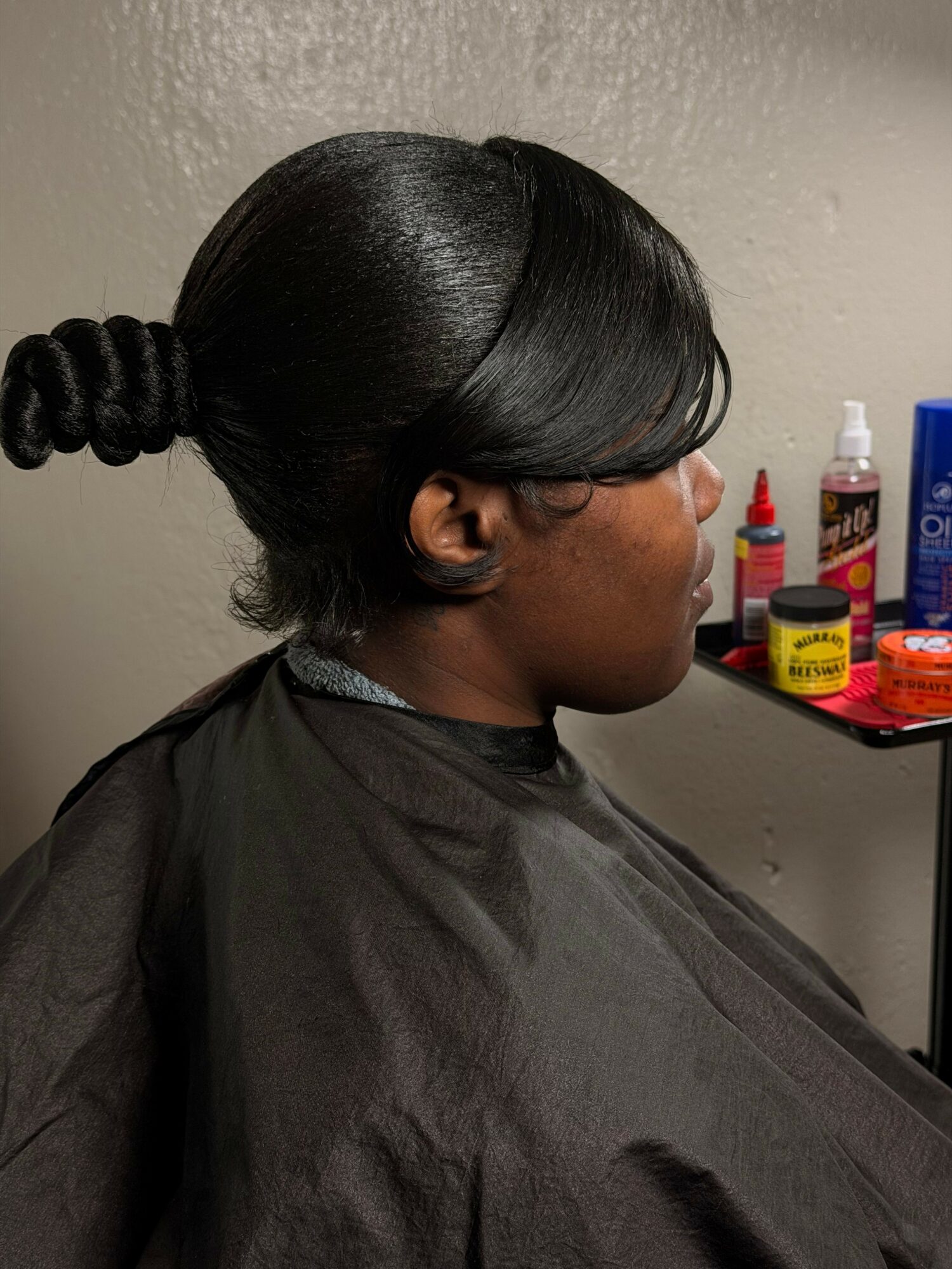 Side view of a person with shiny, styled hair sitting in a salon chair, facing a mirror, with hair products on a shelf behind.