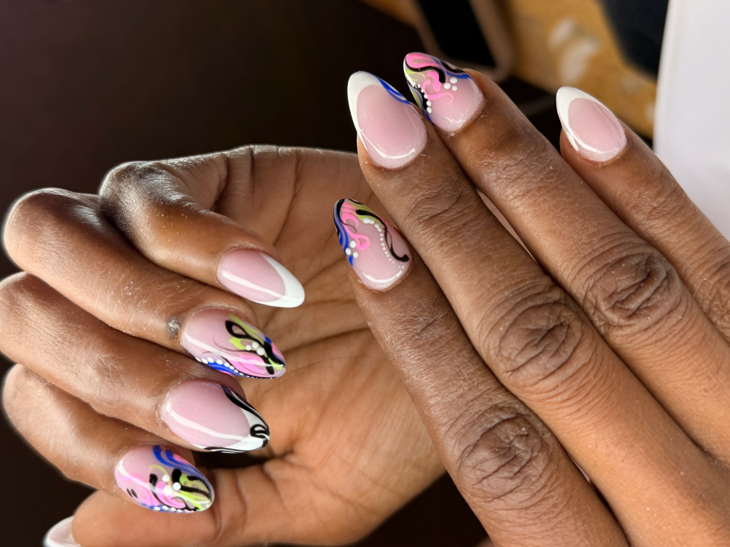 Close-up of hands with manicured nails featuring pink, black, blue, and yellow abstract designs on some nails.