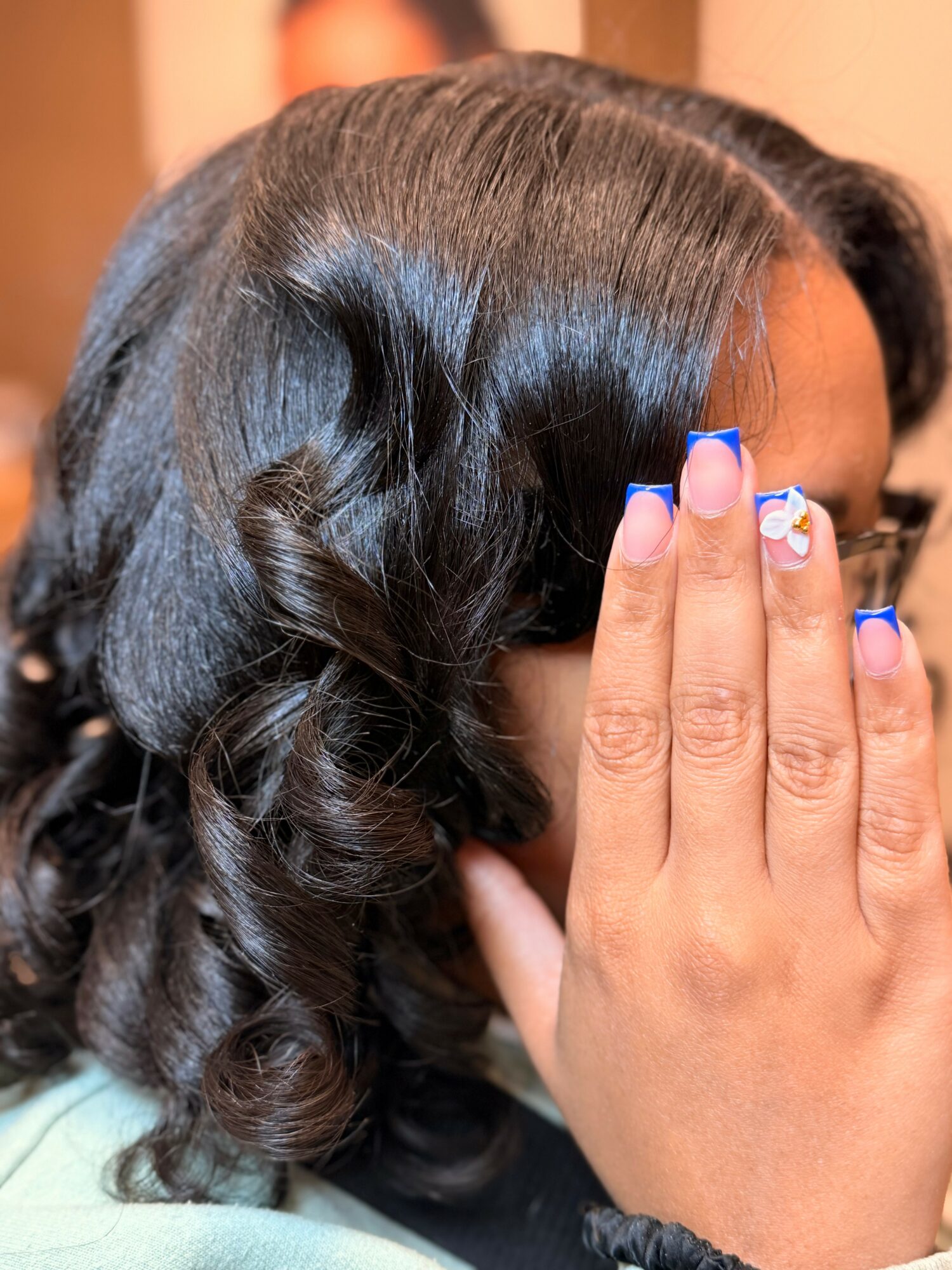 Person with dark, curly hair covering face, hand with decorated nails near face, background blurred.