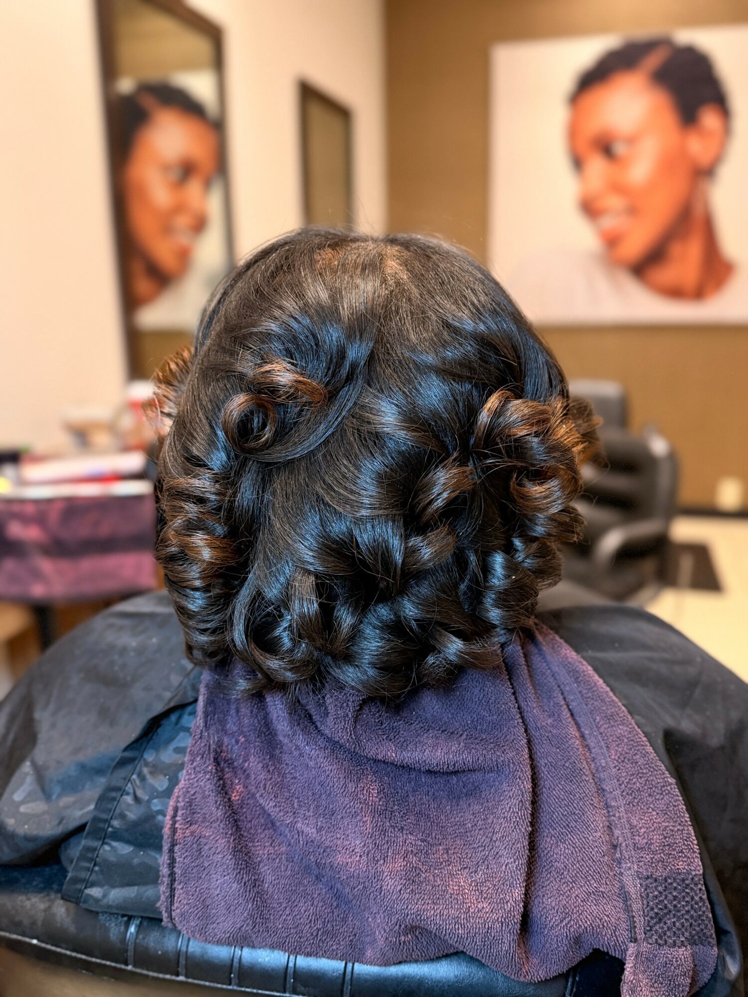 Back of a person's head with styled, shiny, dark hair in loose curls, sitting in a salon chair with portraits on the wall.