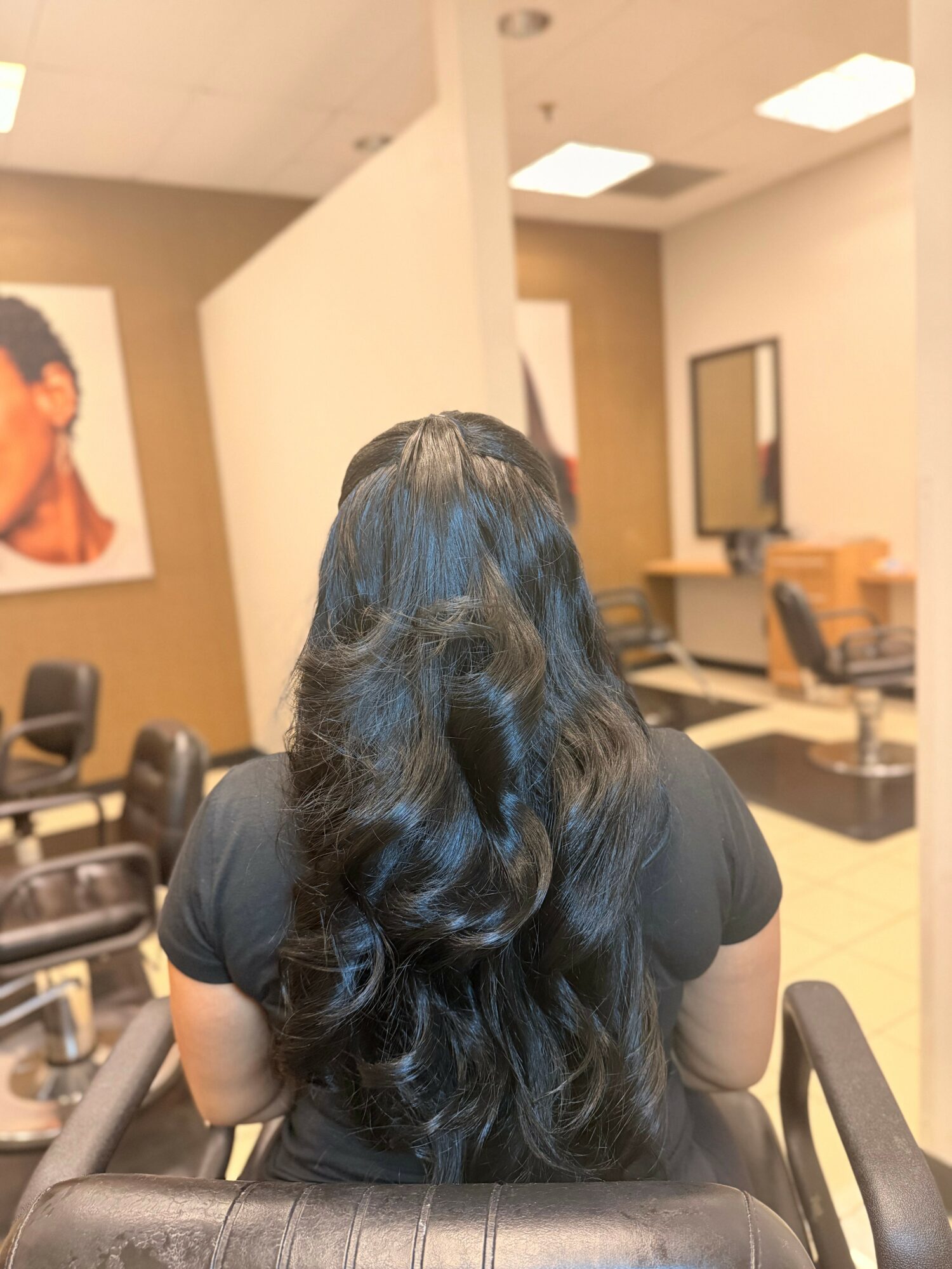 Back view of a woman with long, wavy black hair sitting in a salon chair, facing away from the camera.