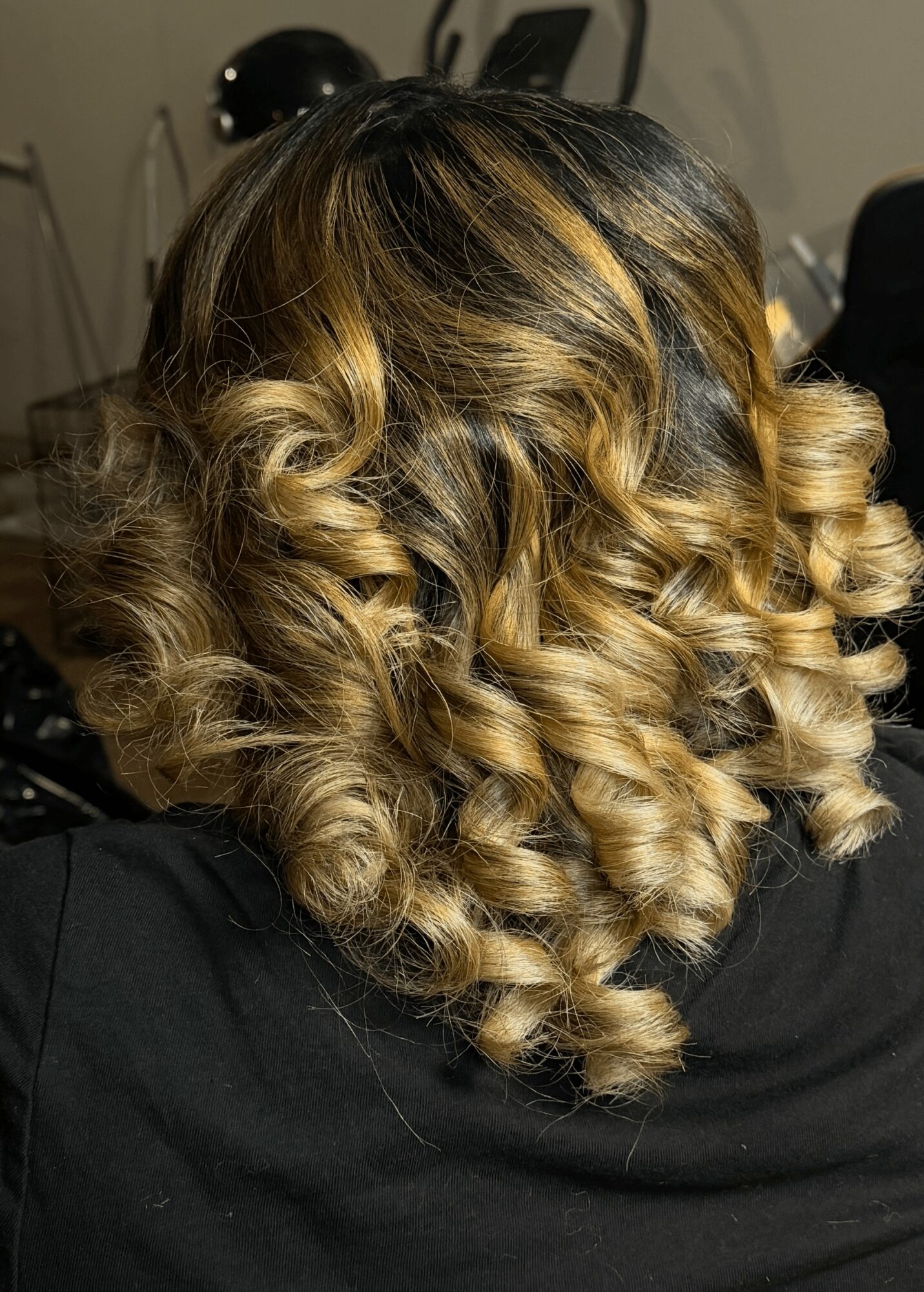 Back view of a woman with shoulder-length, curly hair with blonde highlights, wearing a black top.