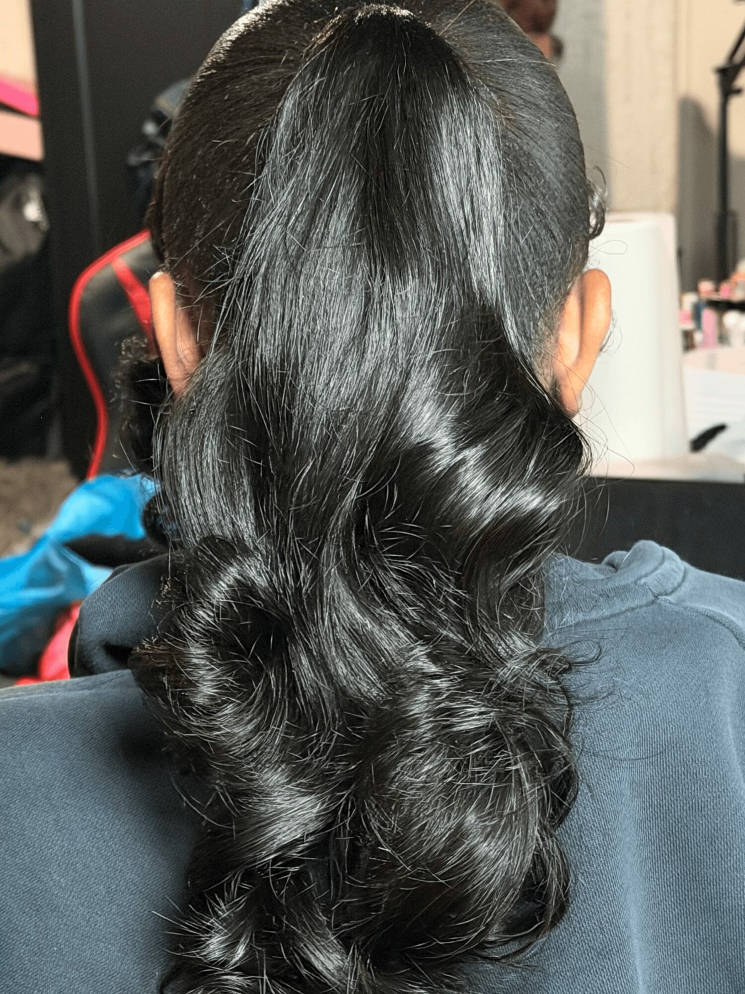 Back of person's head with long, wavy black hair, sitting indoors, wearing a dark top.