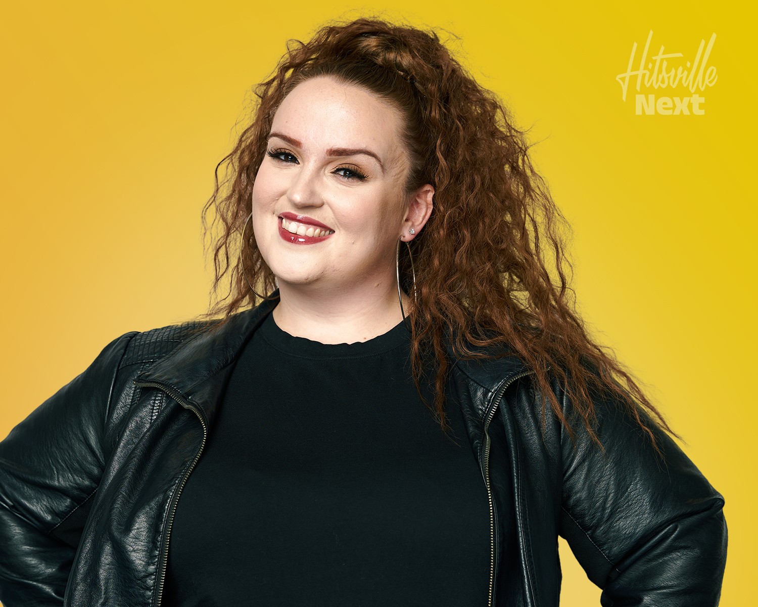 Woman with curly hair smiling, wearing a black top and leather jacket, against yellow background.